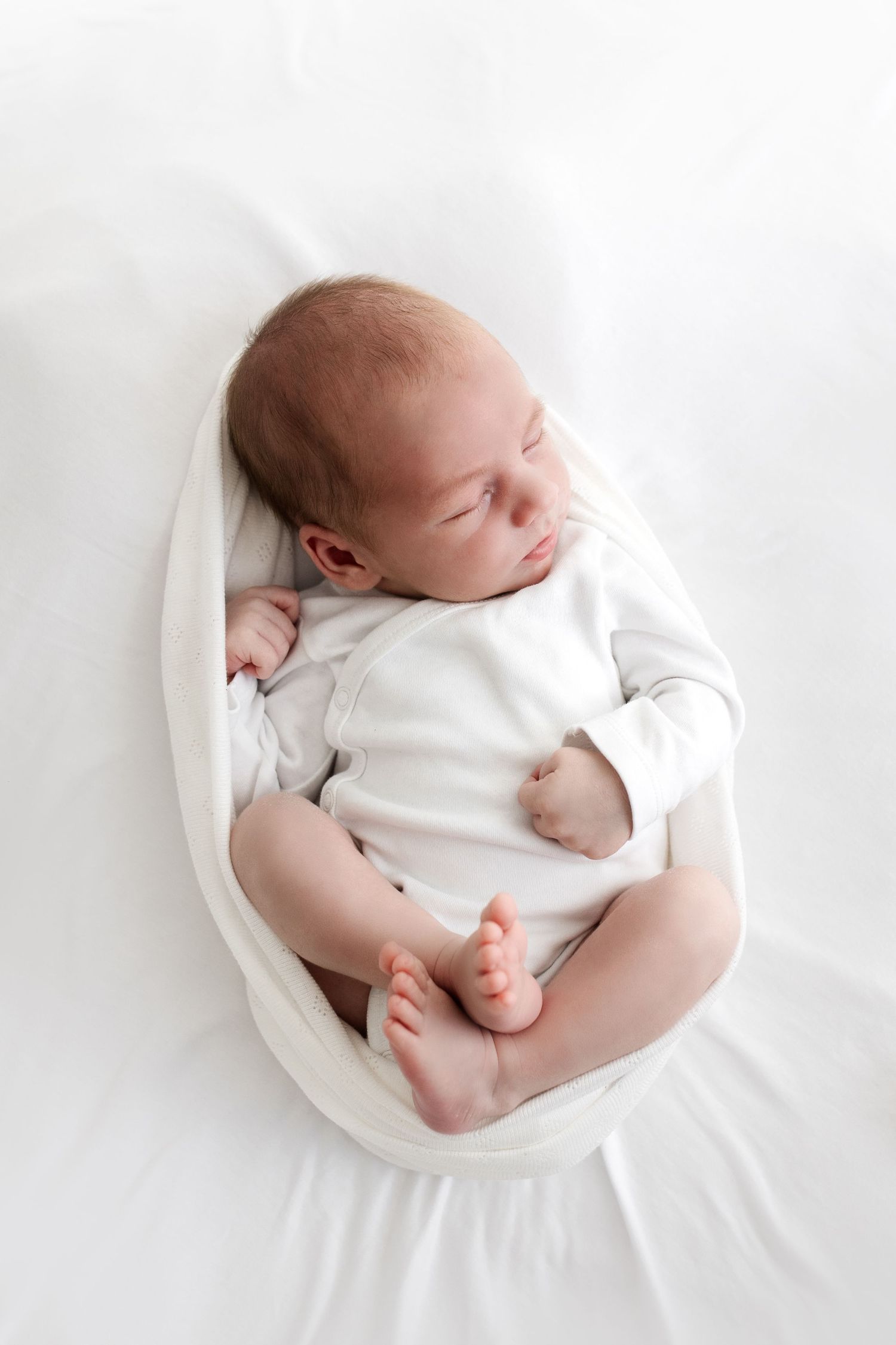 A peaceful newborn baby in a white onesie sleeps curled up on a soft white blanket.