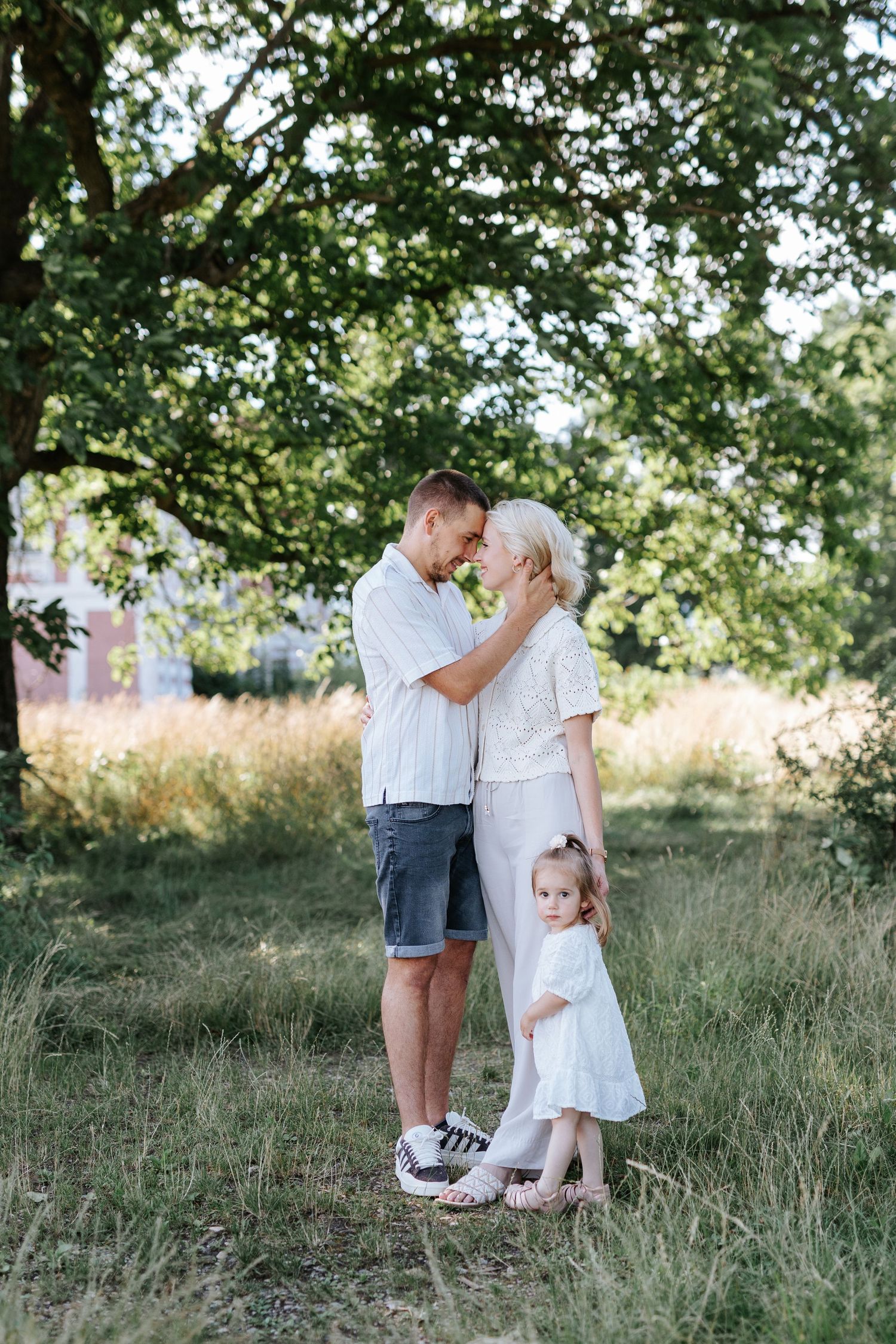 Couple touching foreheads tenderly with young daughter beside them under a large tree outdoors.