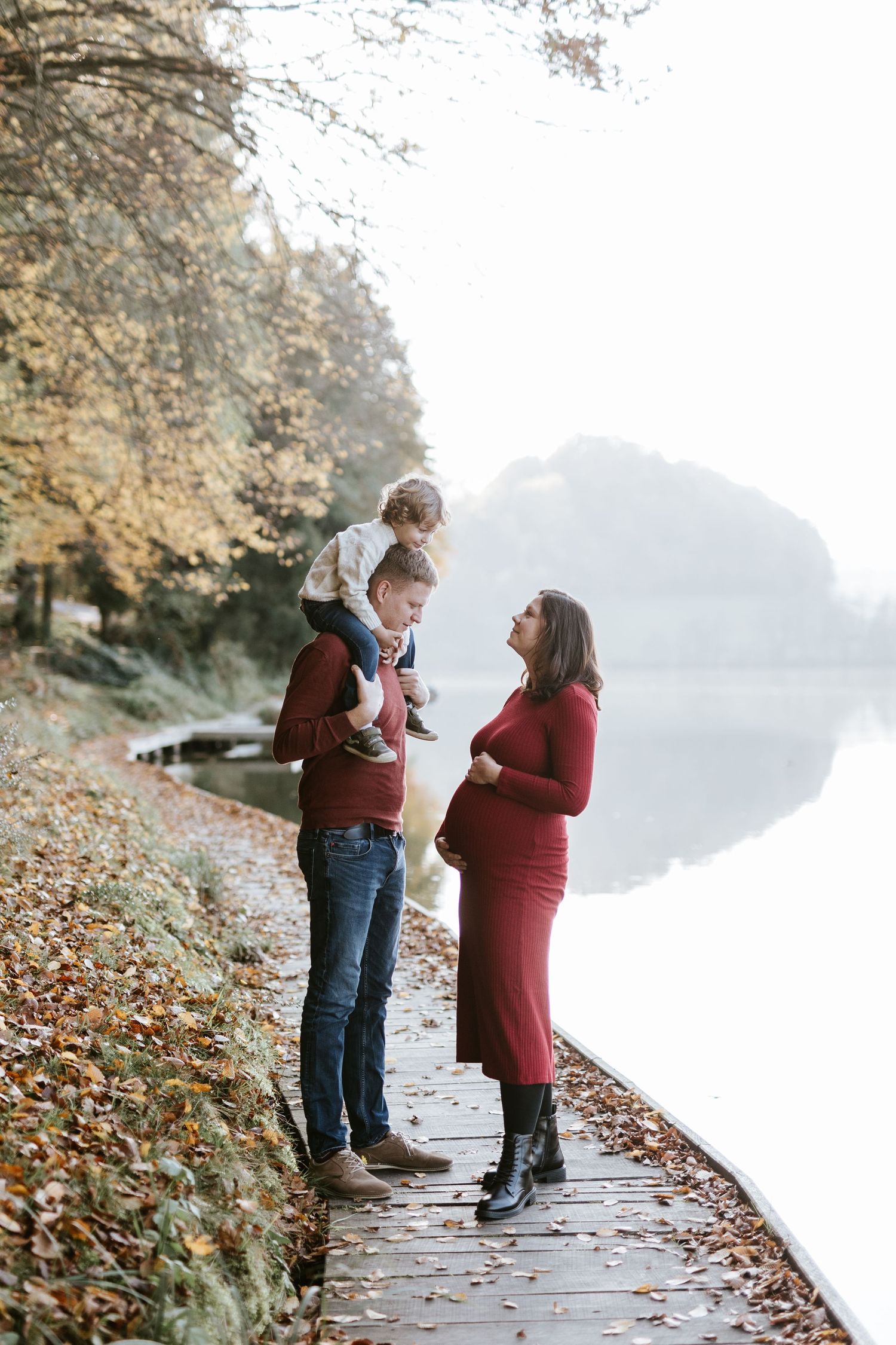 Pregnant woman in red dress with family on a misty autumn lakeside boardwalk.