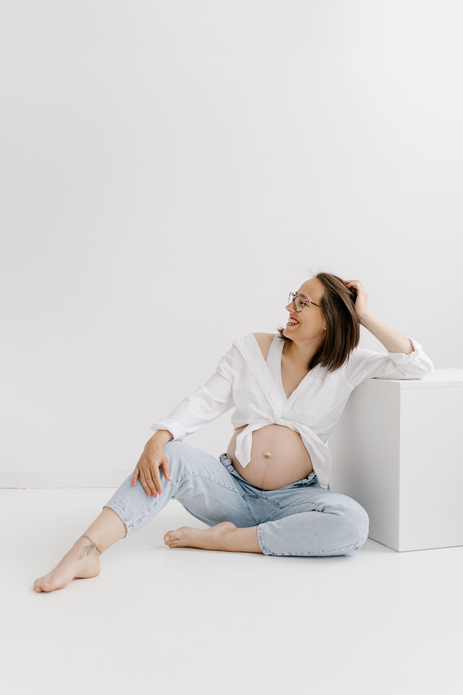 Pregnant woman in glasses and white shirt sitting on floor leaning on white cube looking away.