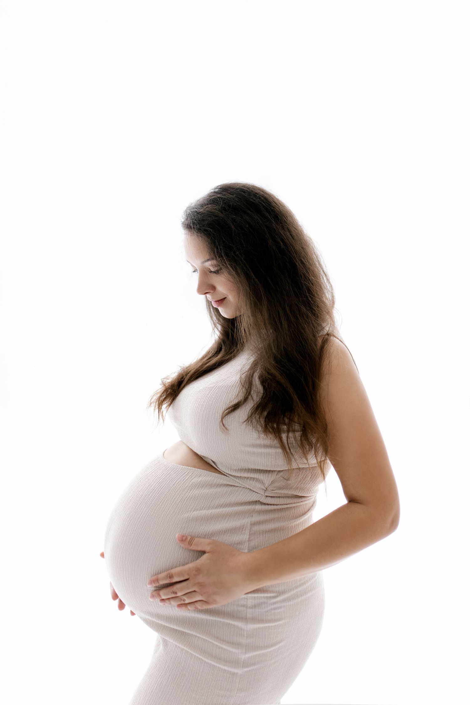 Pregnant woman in beige dress holding baby bump against white background.