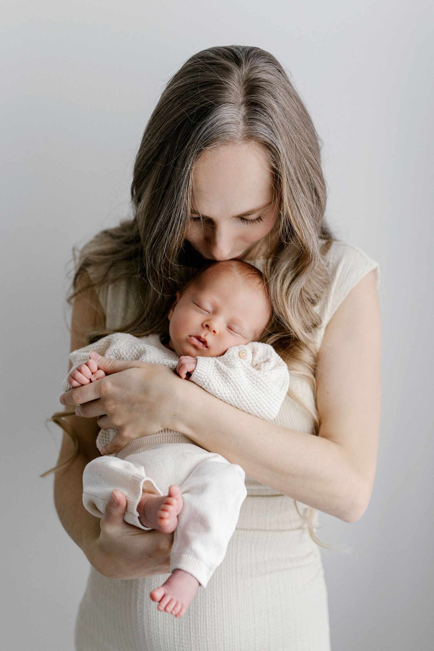 Mother in cream dress tenderly kissing sleeping newborn baby held in her arms.