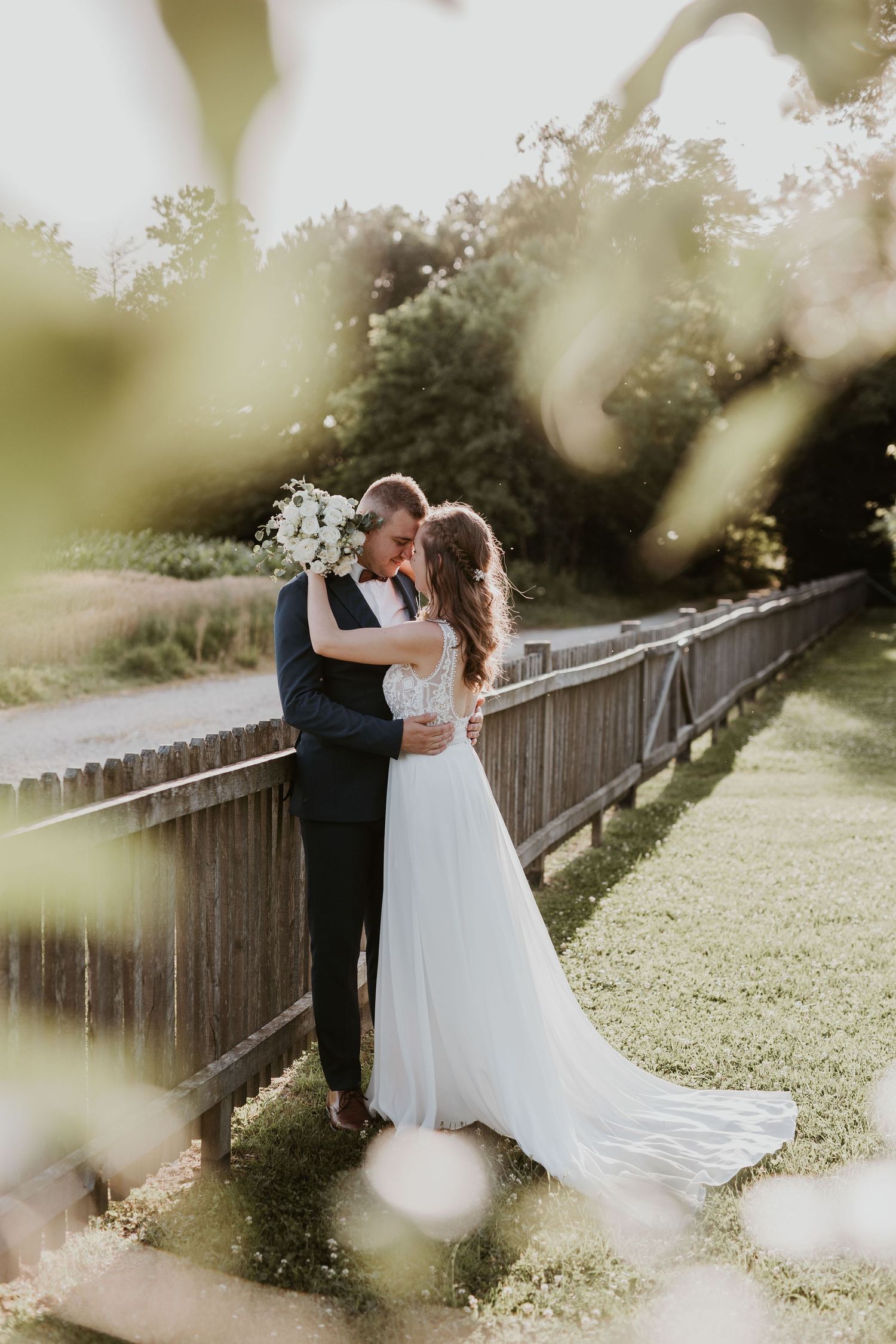 Bride and groom embrace tenderly at sunset near a wooden fence, bride in lace backless gown.