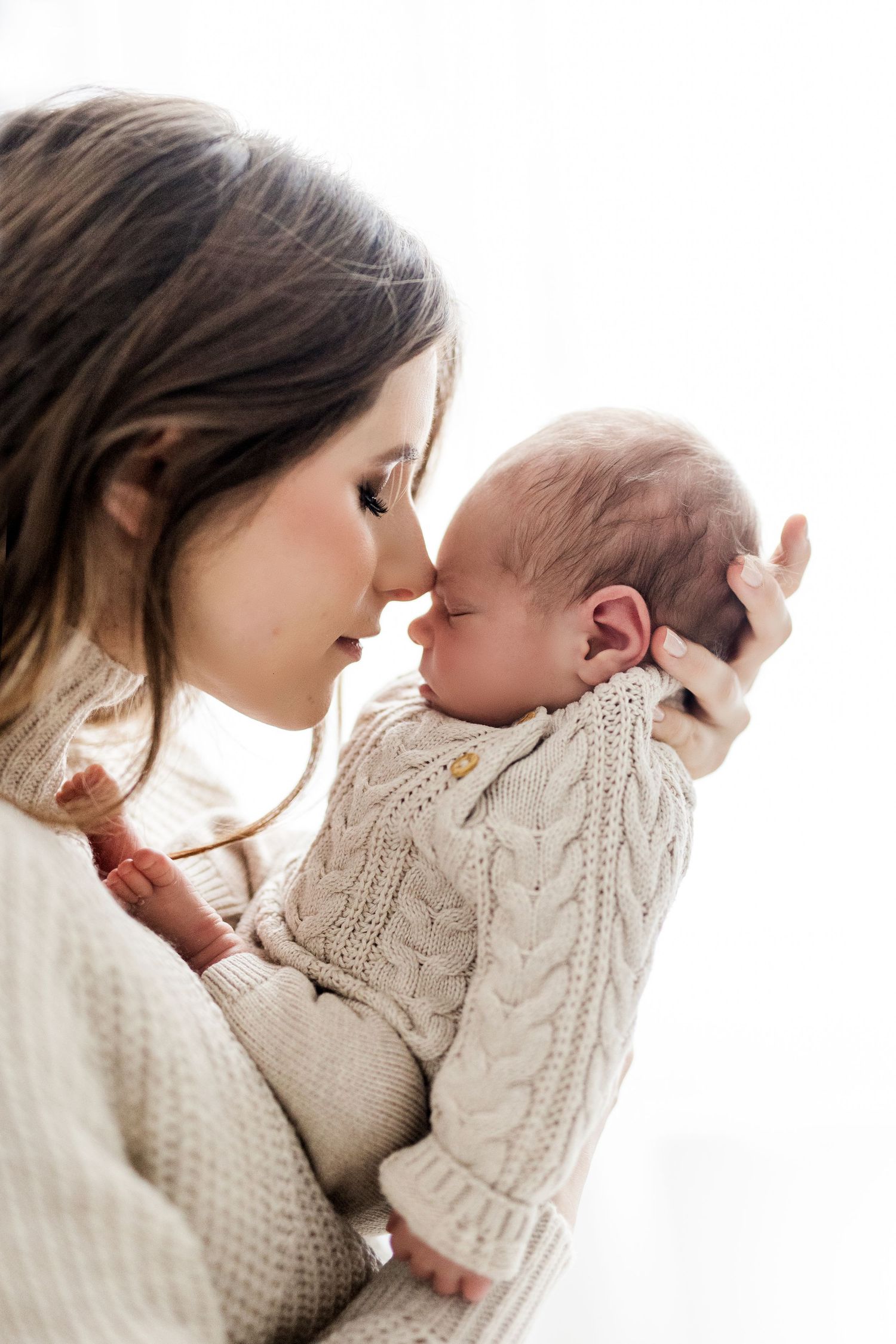 A mother tenderly touches foreheads with her sleeping newborn baby, both dressed in cream knit sweaters.