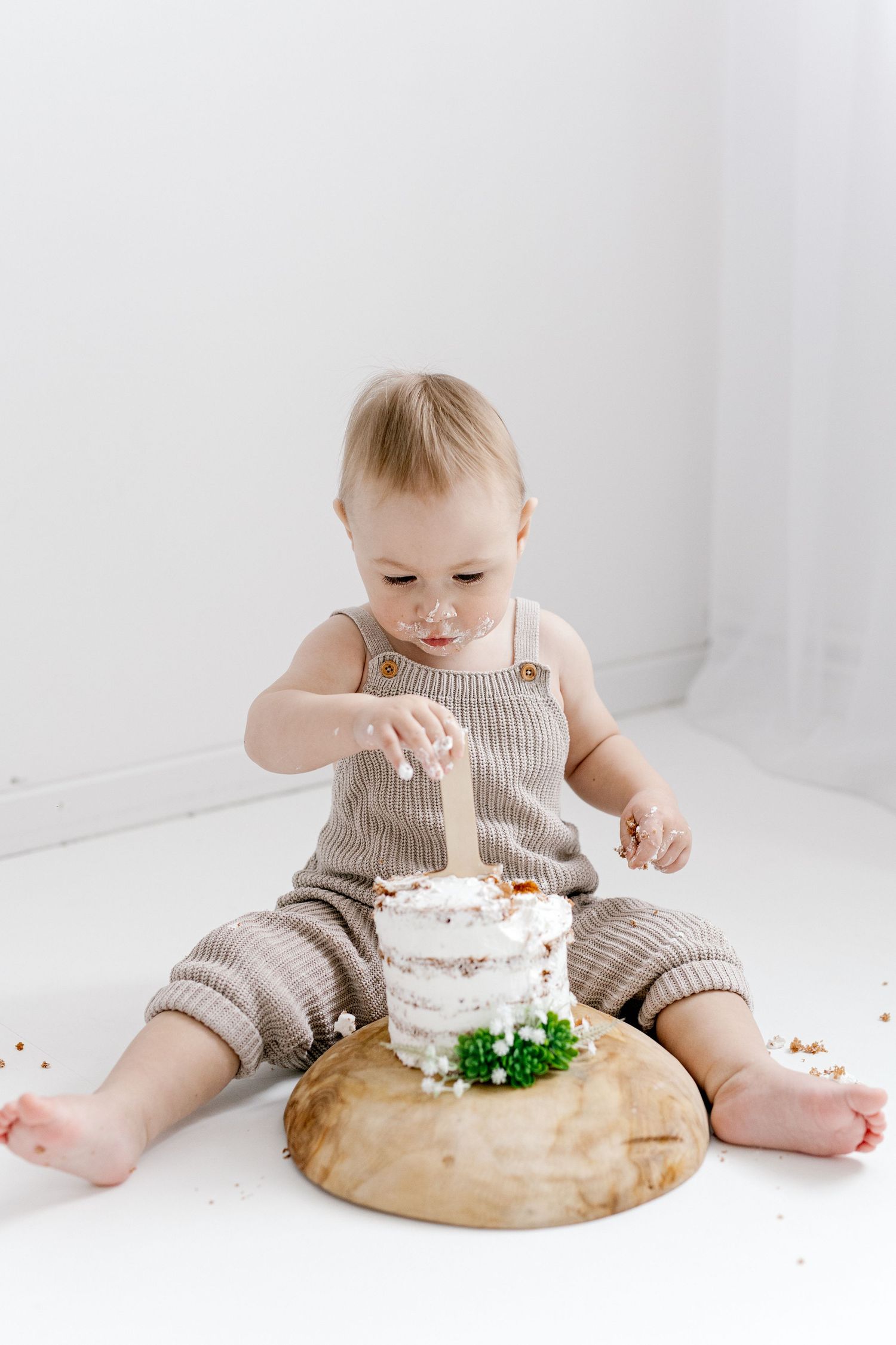 Baby in knit romper sitting and touching first birthday cake smash on wooden board.