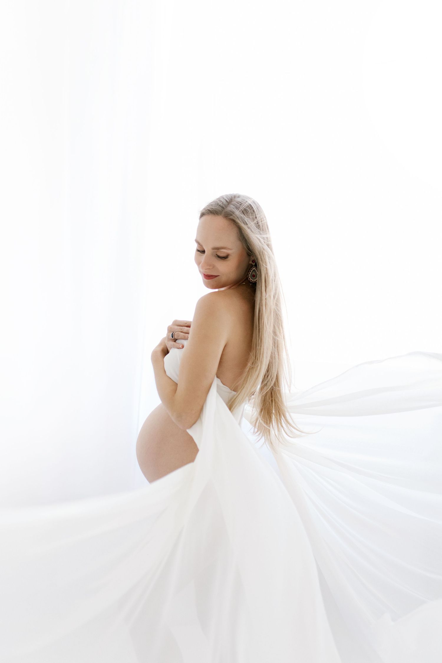Elegantna nosečniška fotografija: nosečnica v padajoči beli tkanini v studiu.