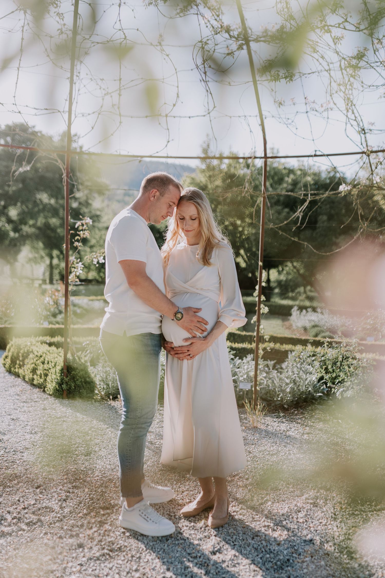 Expecting couple tenderly cradling baby bump in sunlit garden, man in white tee embracing woman in cream dress.