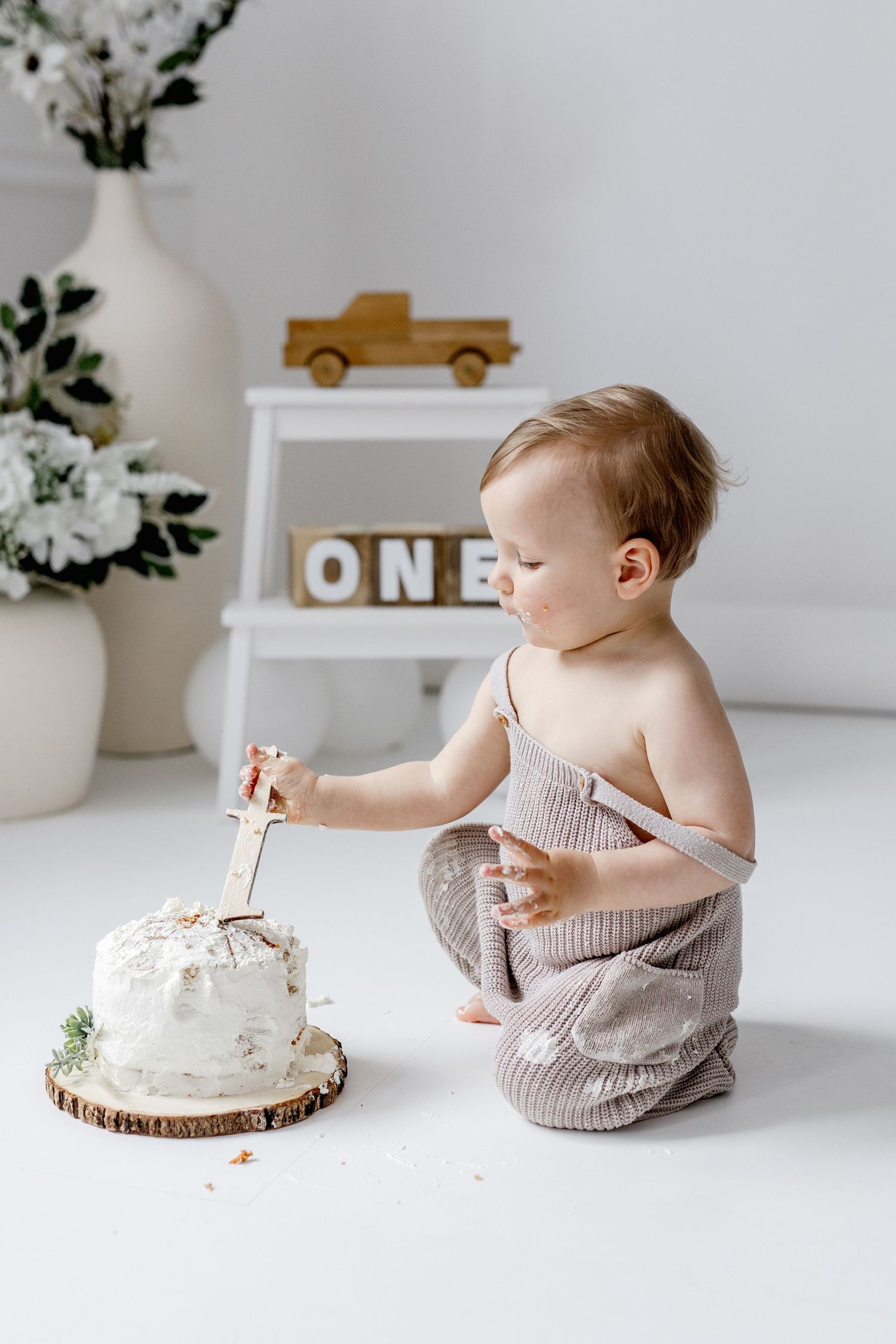 Baby boy in knit romper poking first birthday smash cake with wooden knife.