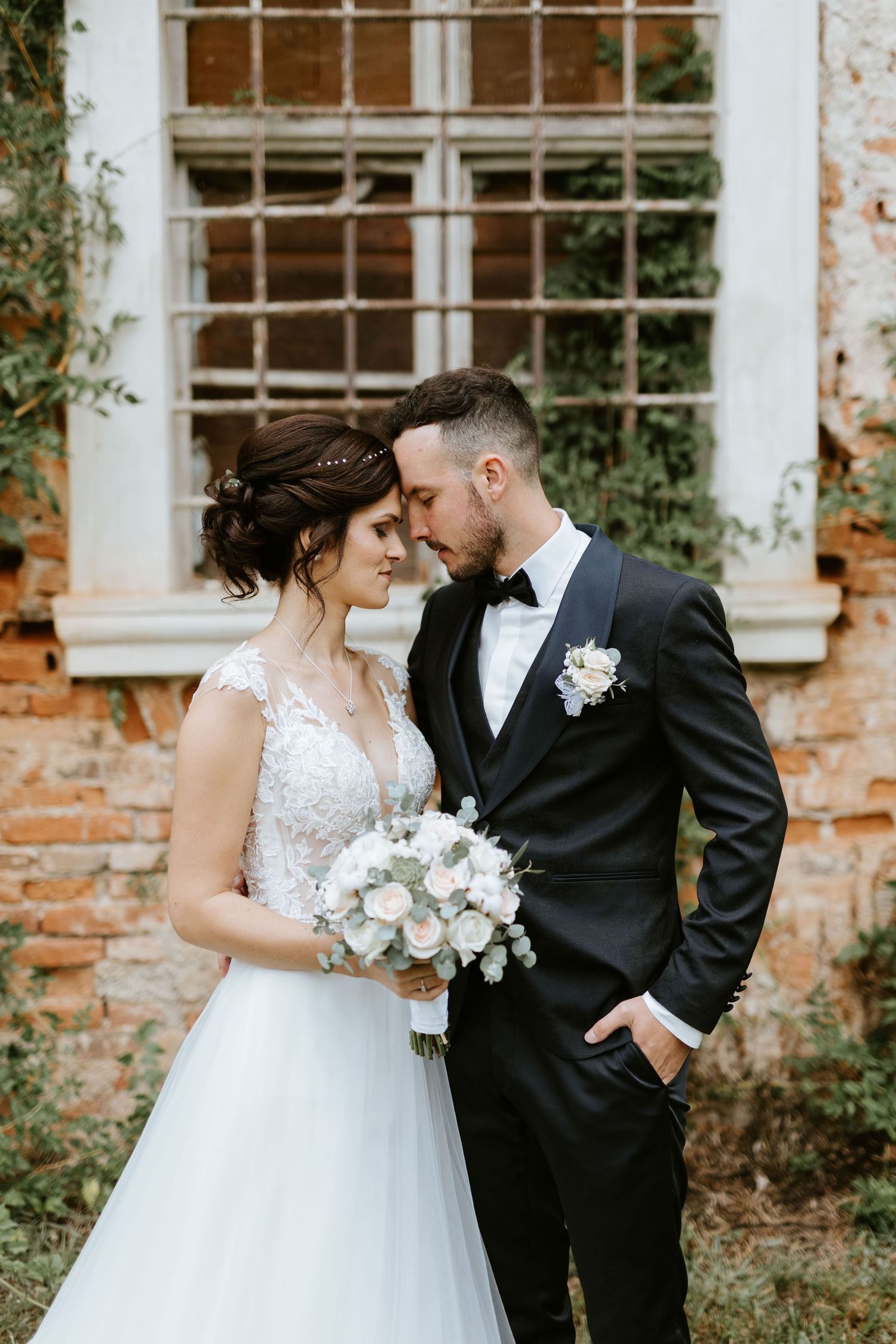 Bride and groom in romantic embrace before ivy-covered brick wall, groom in black tuxedo with boutonniere.
