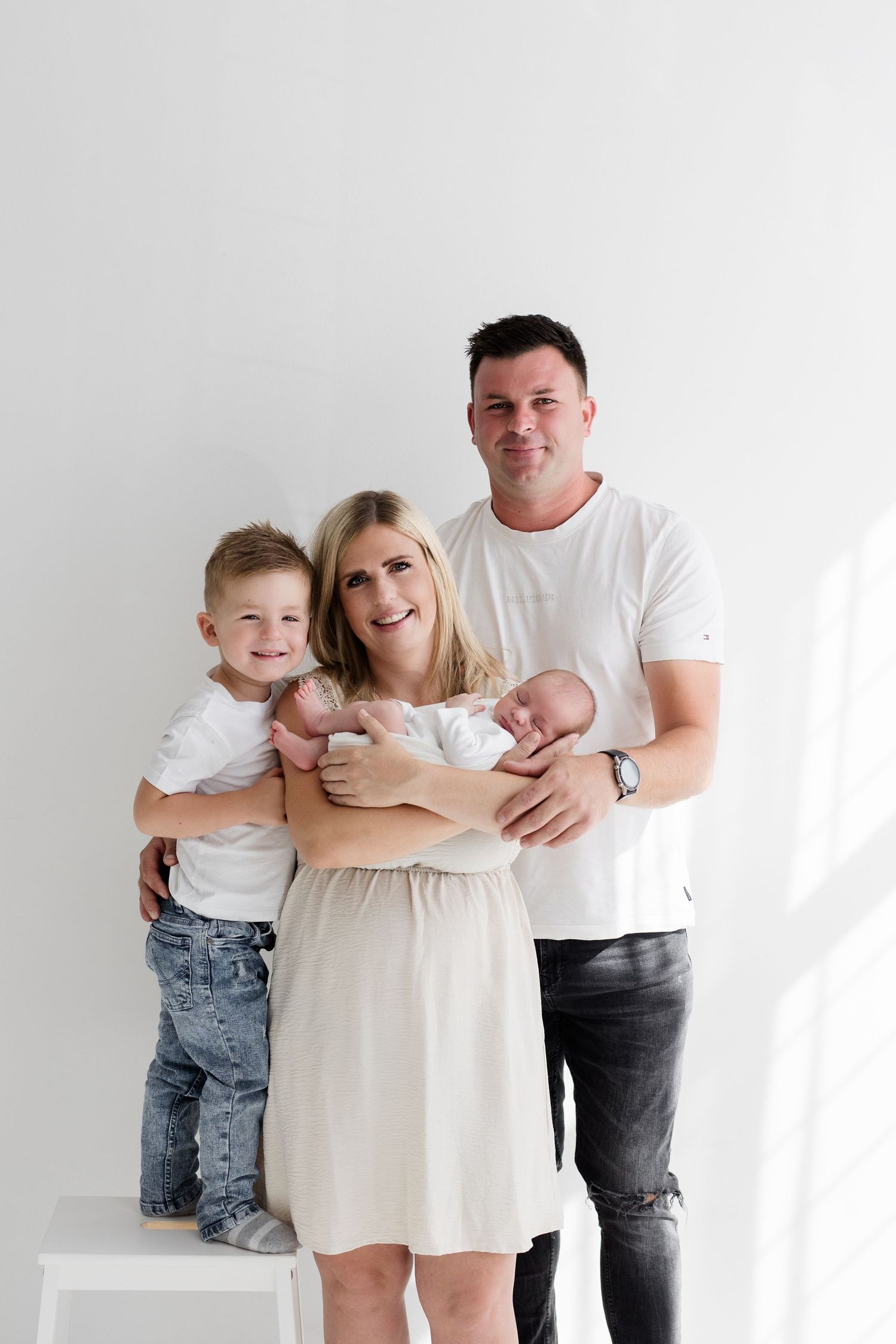 Happy family portrait with parents, young son, and newborn twins dressed in white against white wall.