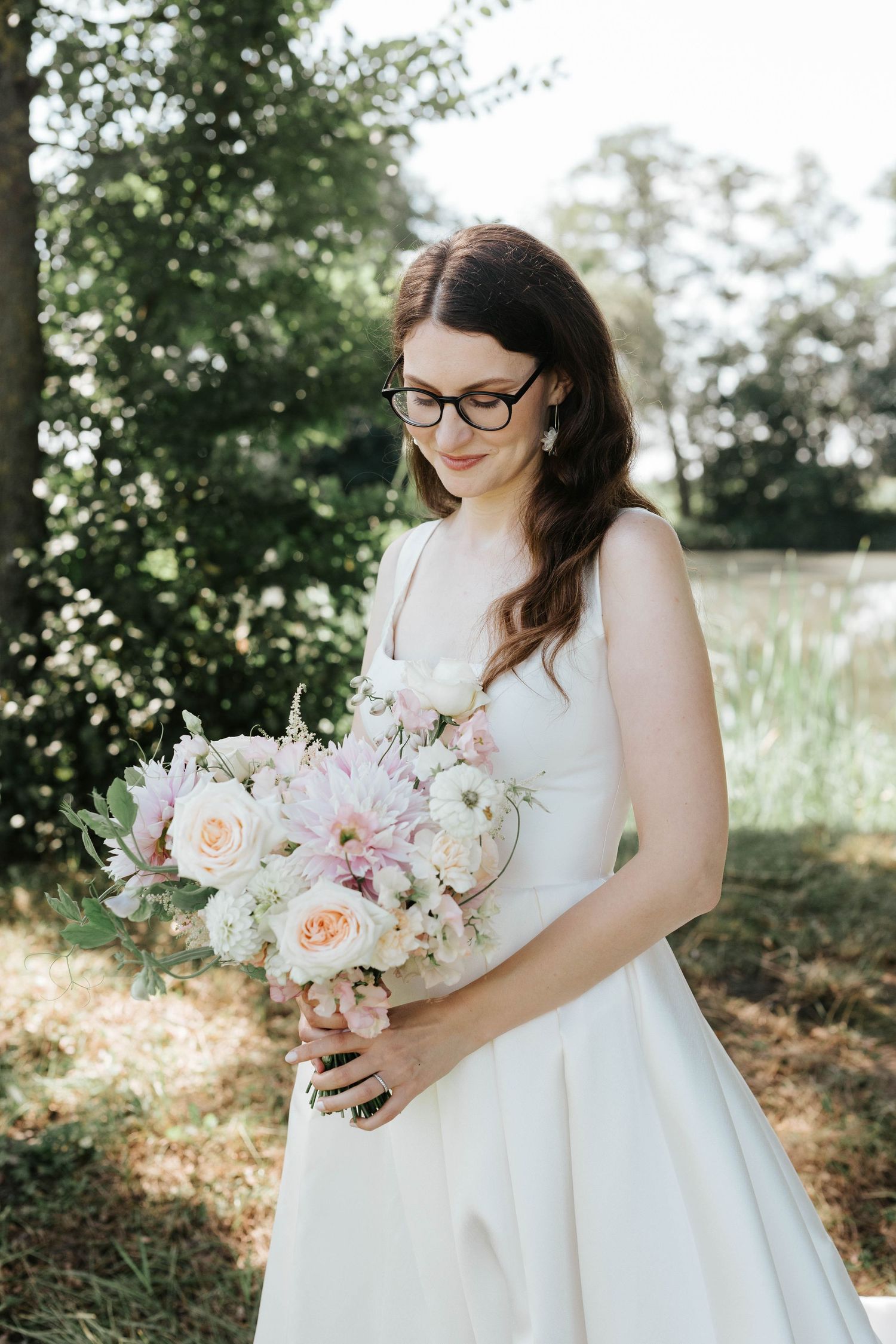 Brunette bride wearing glasses looking down at pastel pink and white bouquet in a sunny garden setting.
