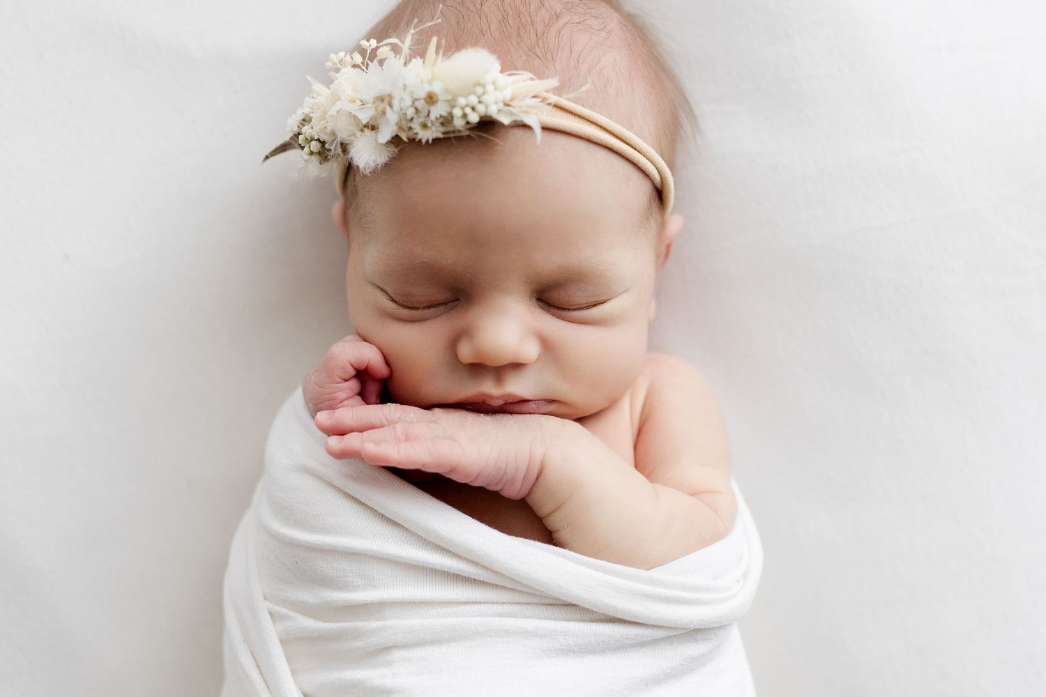 Sleeping newborn baby girl wrapped in white swaddle wearing delicate floral headband.