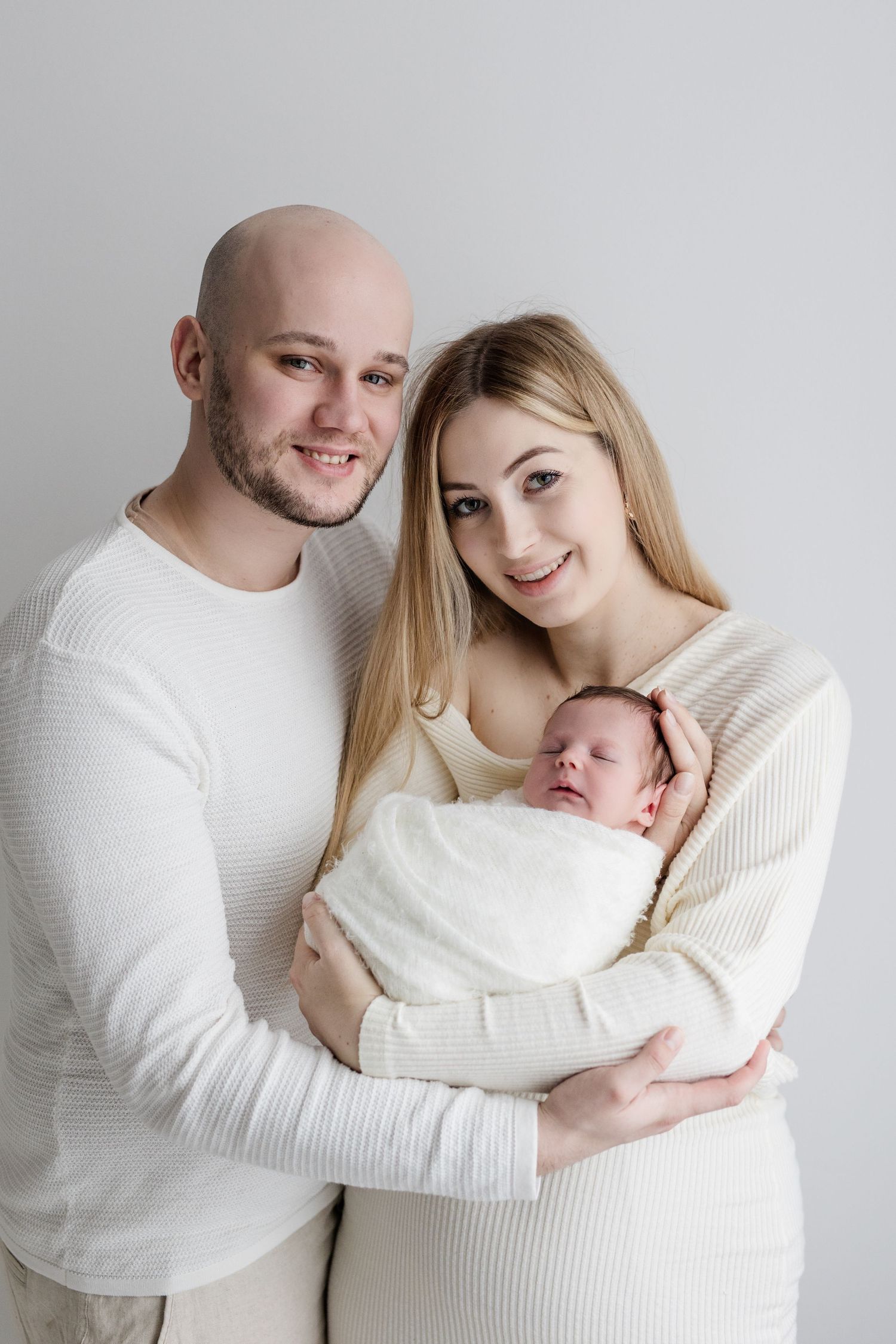 Smiling young family in white outfits pose together holding their swaddled newborn baby in studio portrait.