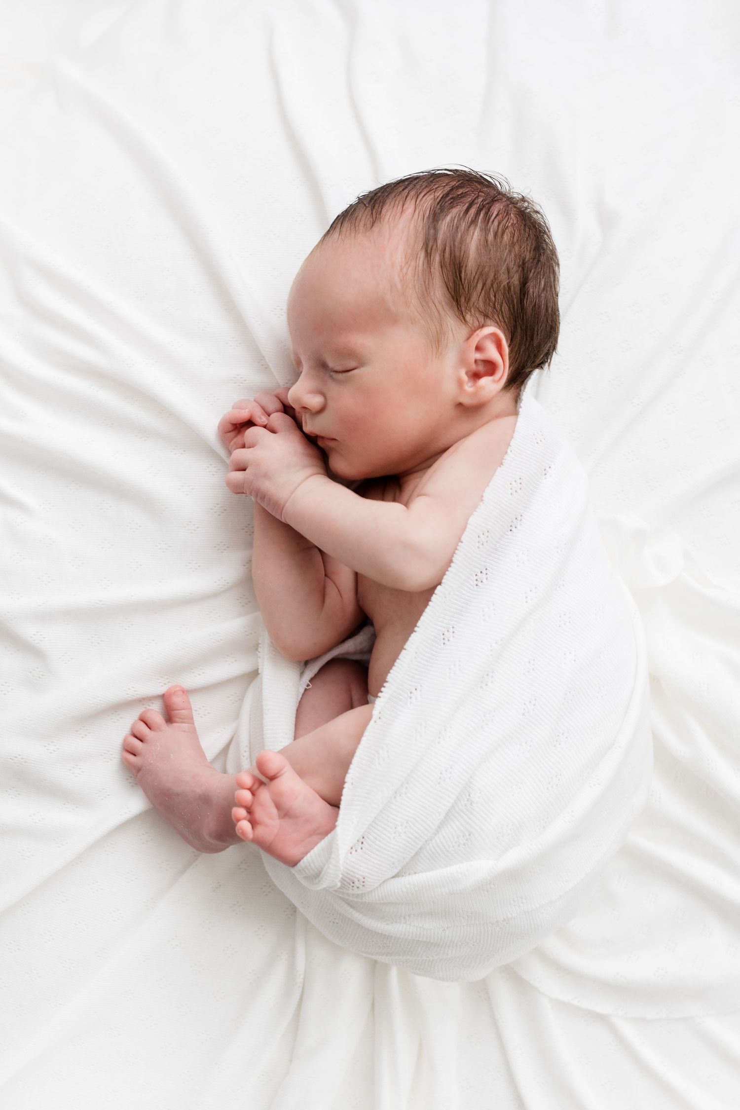 Sleeping newborn baby with dark hair wrapped in white swaddle blanket on white bedding.
