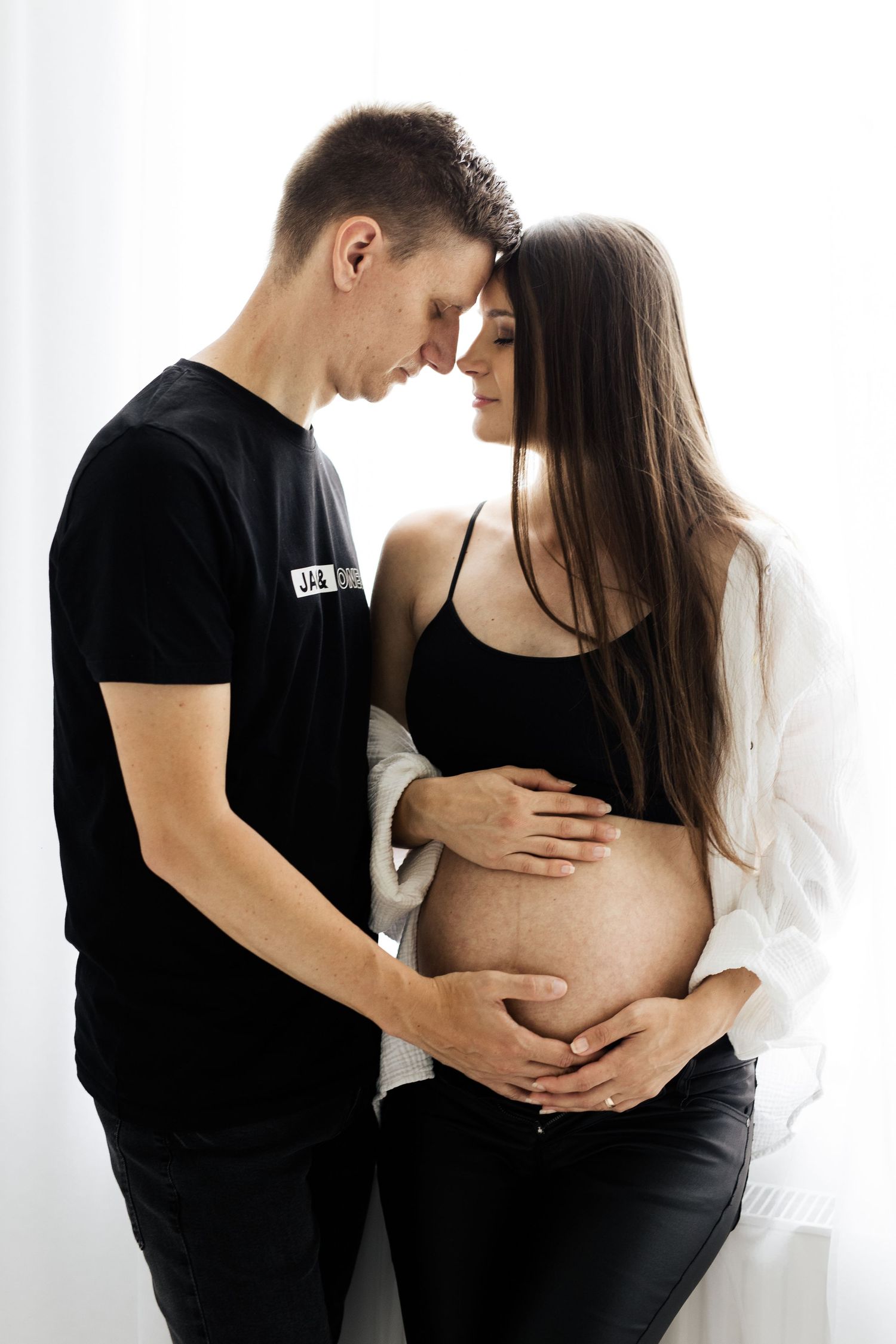Couple touching foreheads tenderly while holding pregnant belly in bright studio.