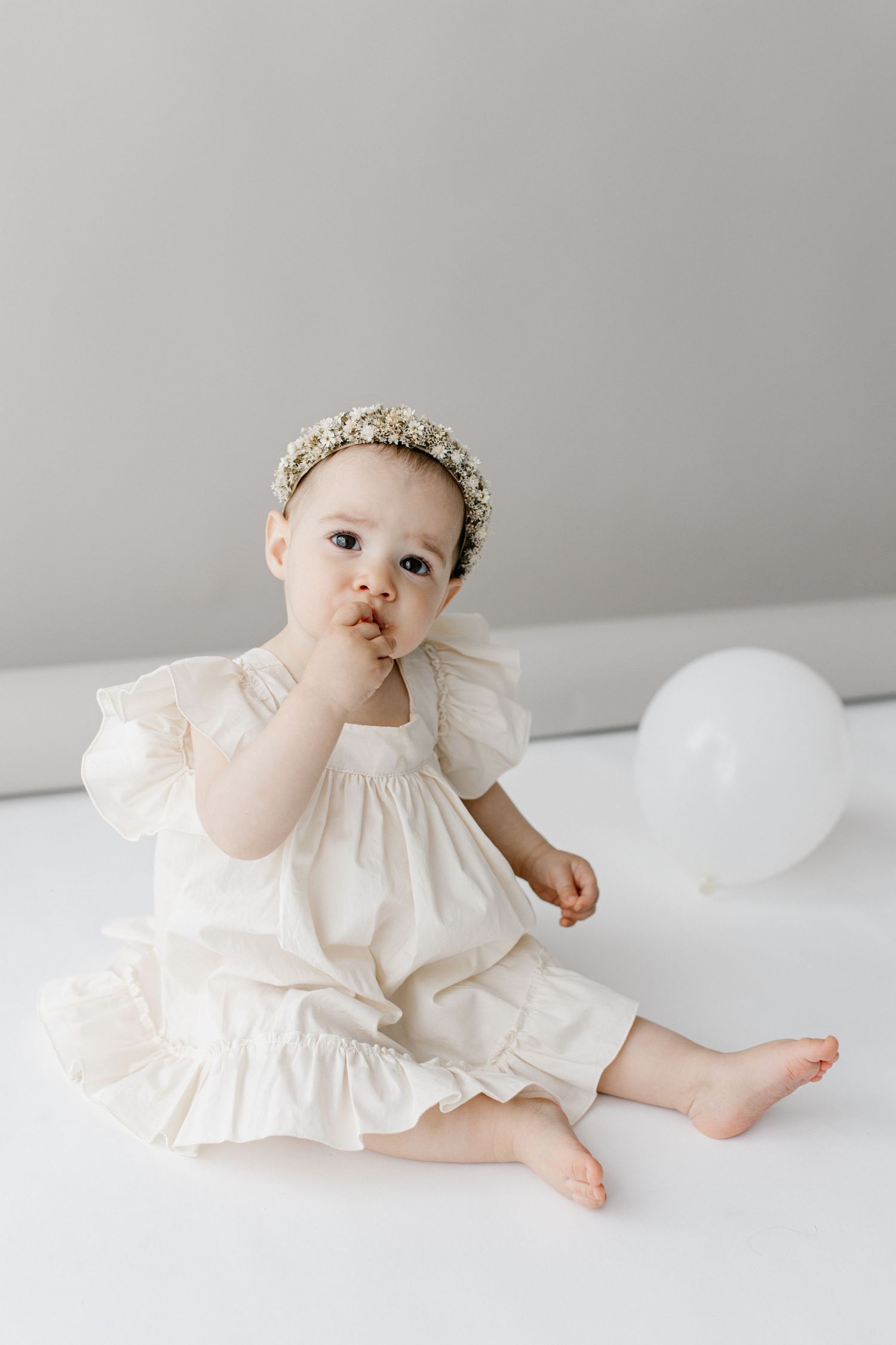 Baby girl in white ruffled dress with floral headband sitting near white balloon.