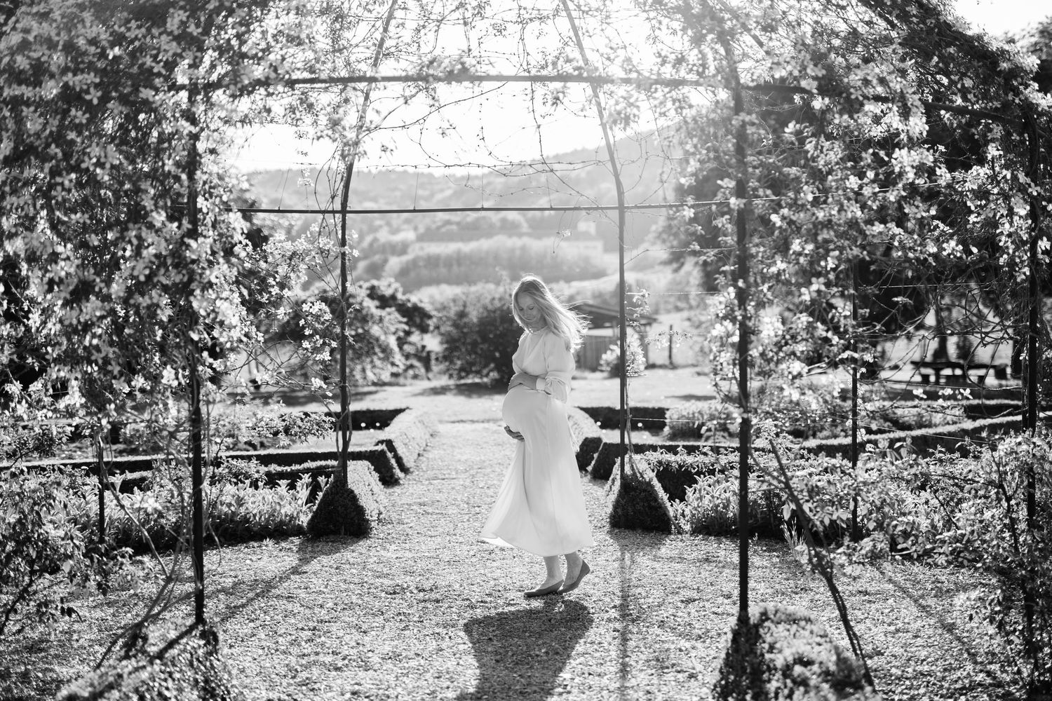 Black and white photo of bride in flowing gown standing under garden arch with scenic countryside backdrop.