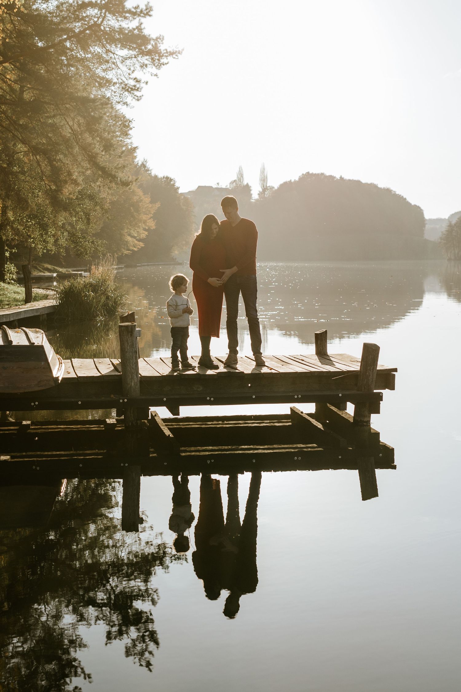 Family of three silhouetted on wooden dock at golden hour with calm lake and island reflection below.