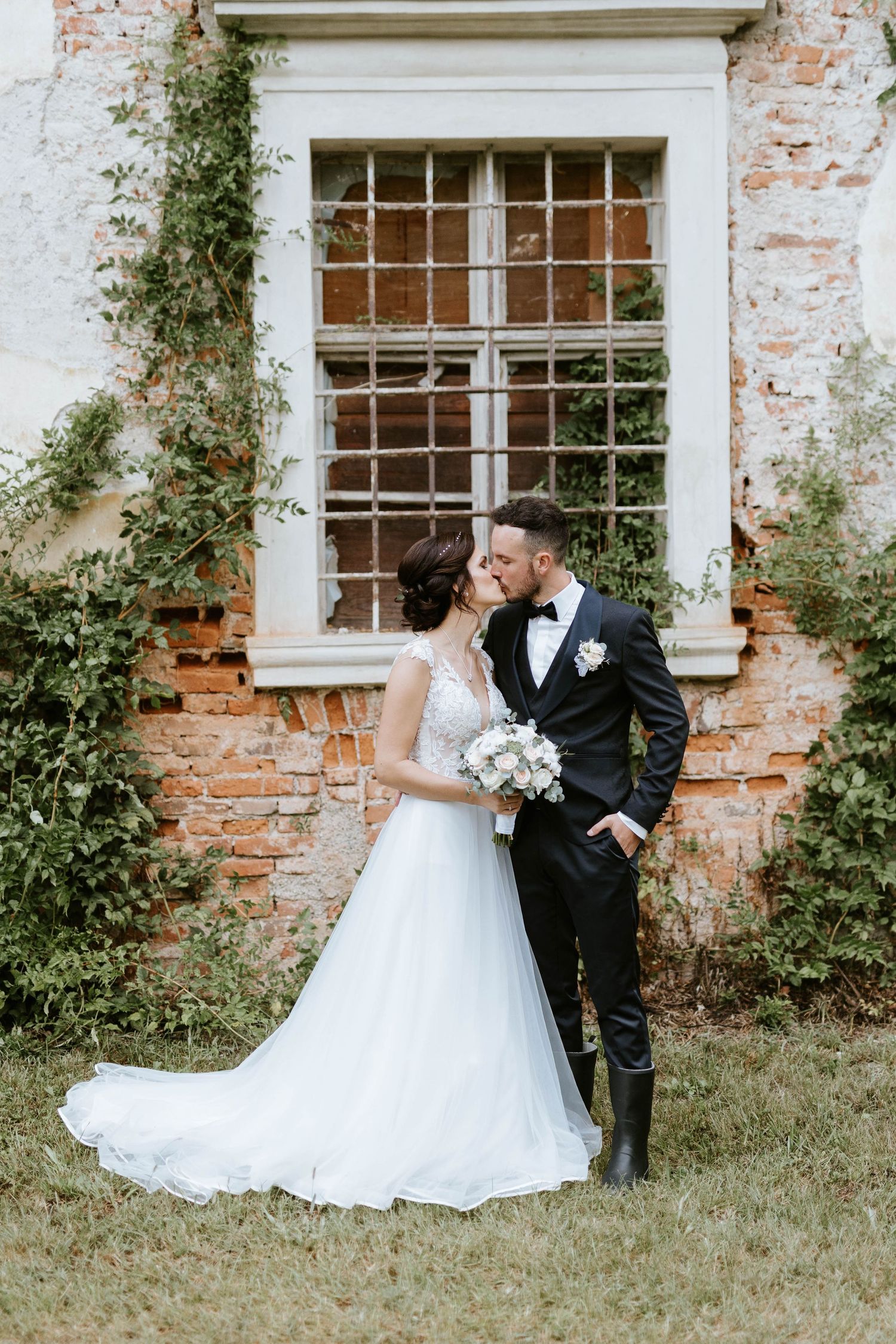 Bride and groom kissing in front of a rustic brick wall with vintage window, bride holding white bouquet.