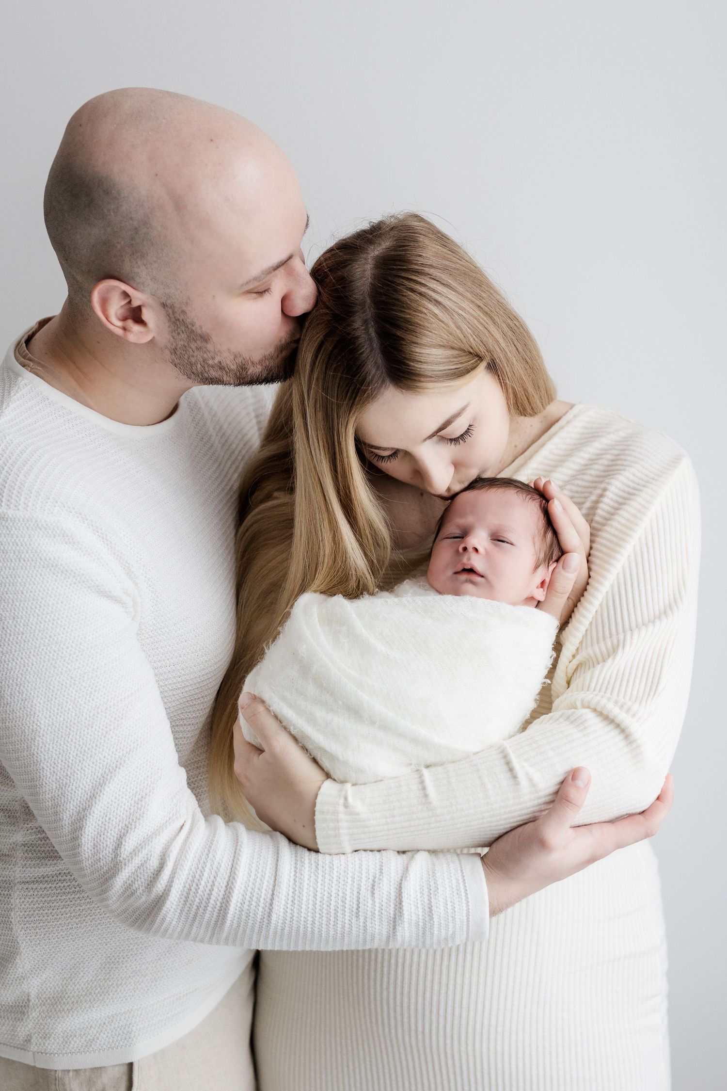 Loving parents in white outfits hold their swaddled newborn baby together in a tender studio portrait.