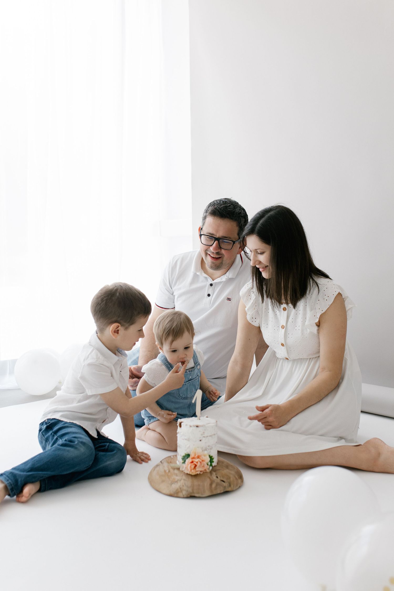 Family of four sitting on white floor celebrating baby's first birthday with small decorated cake.
