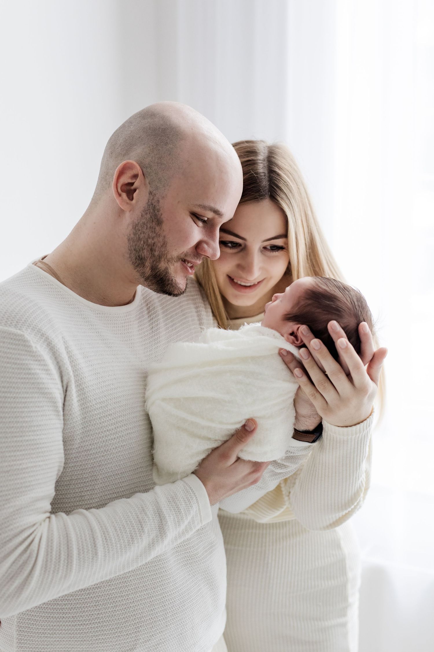 Smiling parents in white outfits tenderly holding and admiring their swaddled newborn baby together.