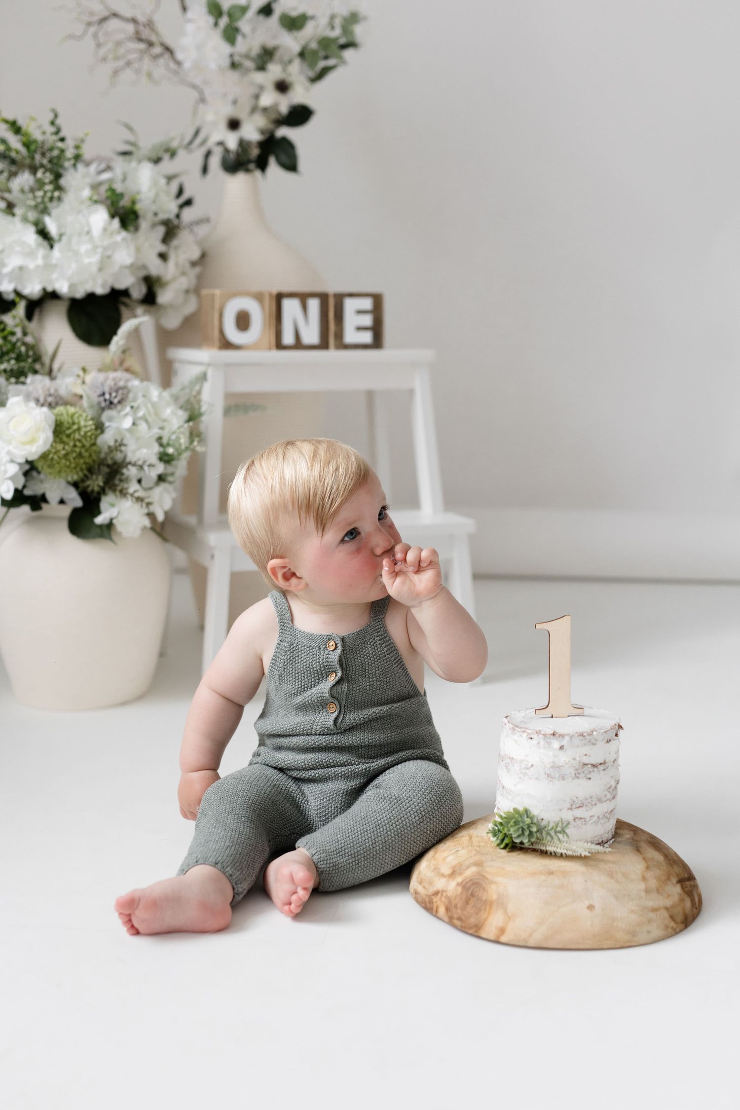 Blond baby in gray romper sitting beside first birthday cake with ONE blocks decor.