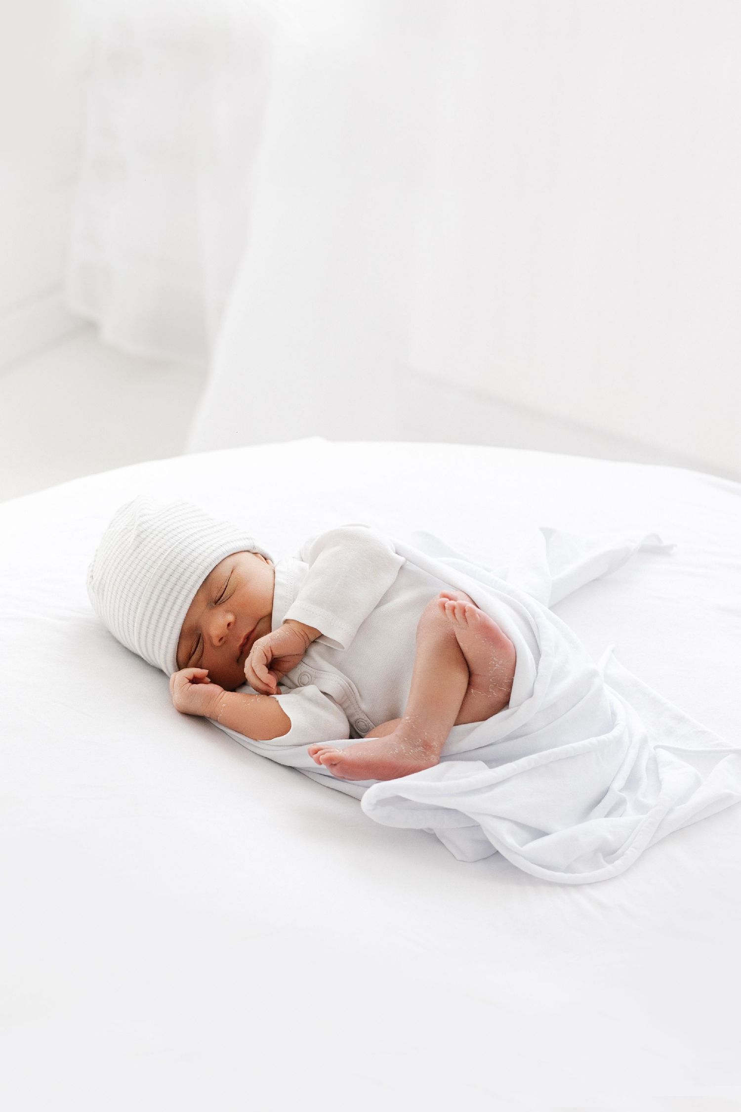 Peaceful newborn baby sleeping on white bed wrapped in soft white blanket wearing striped hospital hat.