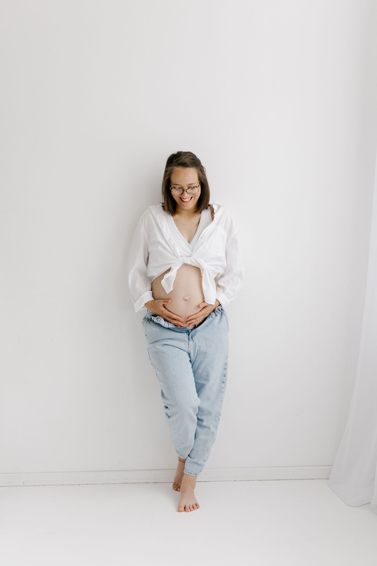 Smiling pregnant woman in glasses and tied white shirt touching her bare baby bump against white wall.