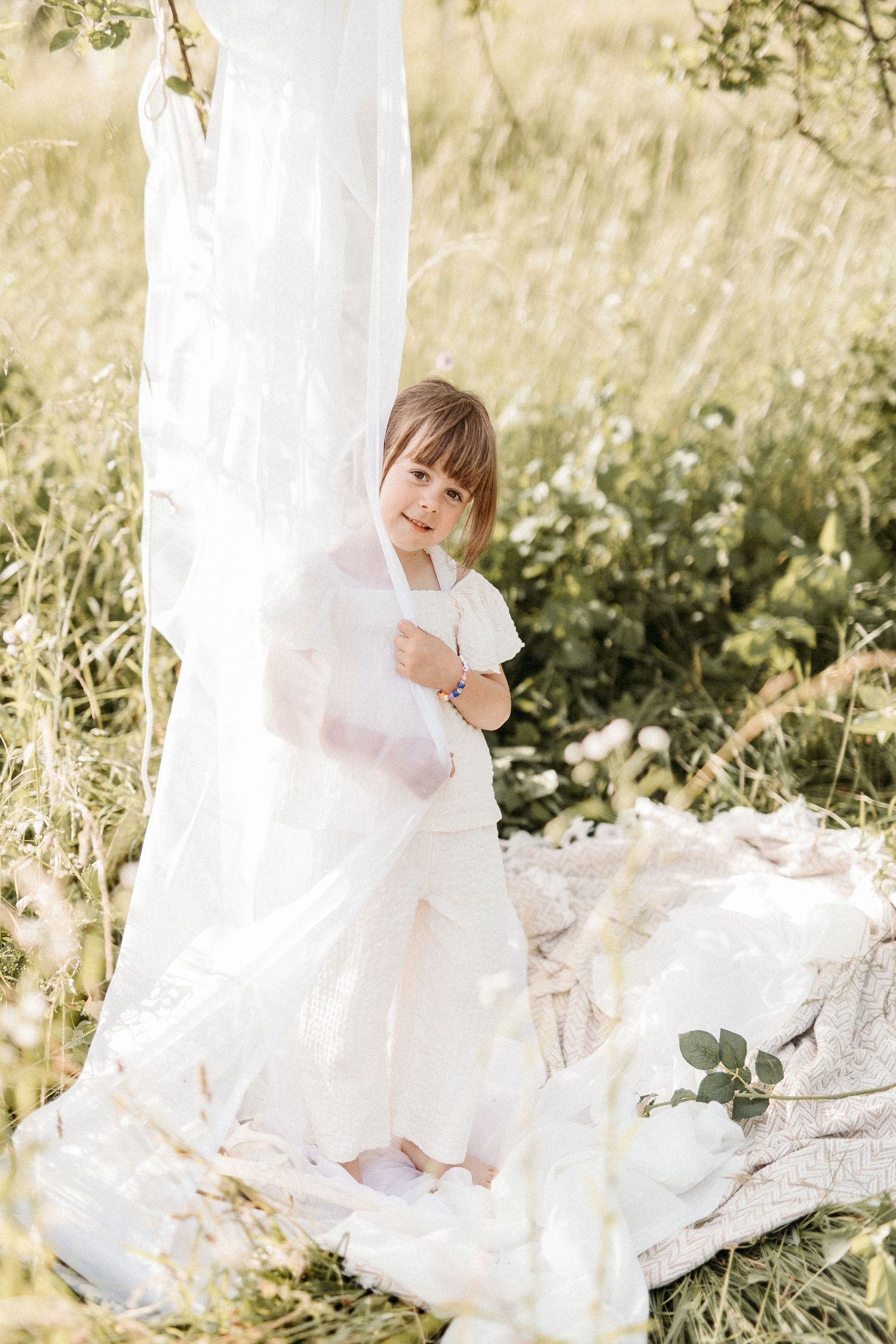 Young girl in white outfit playing with sheer white fabric teepee tent in a sunny field.