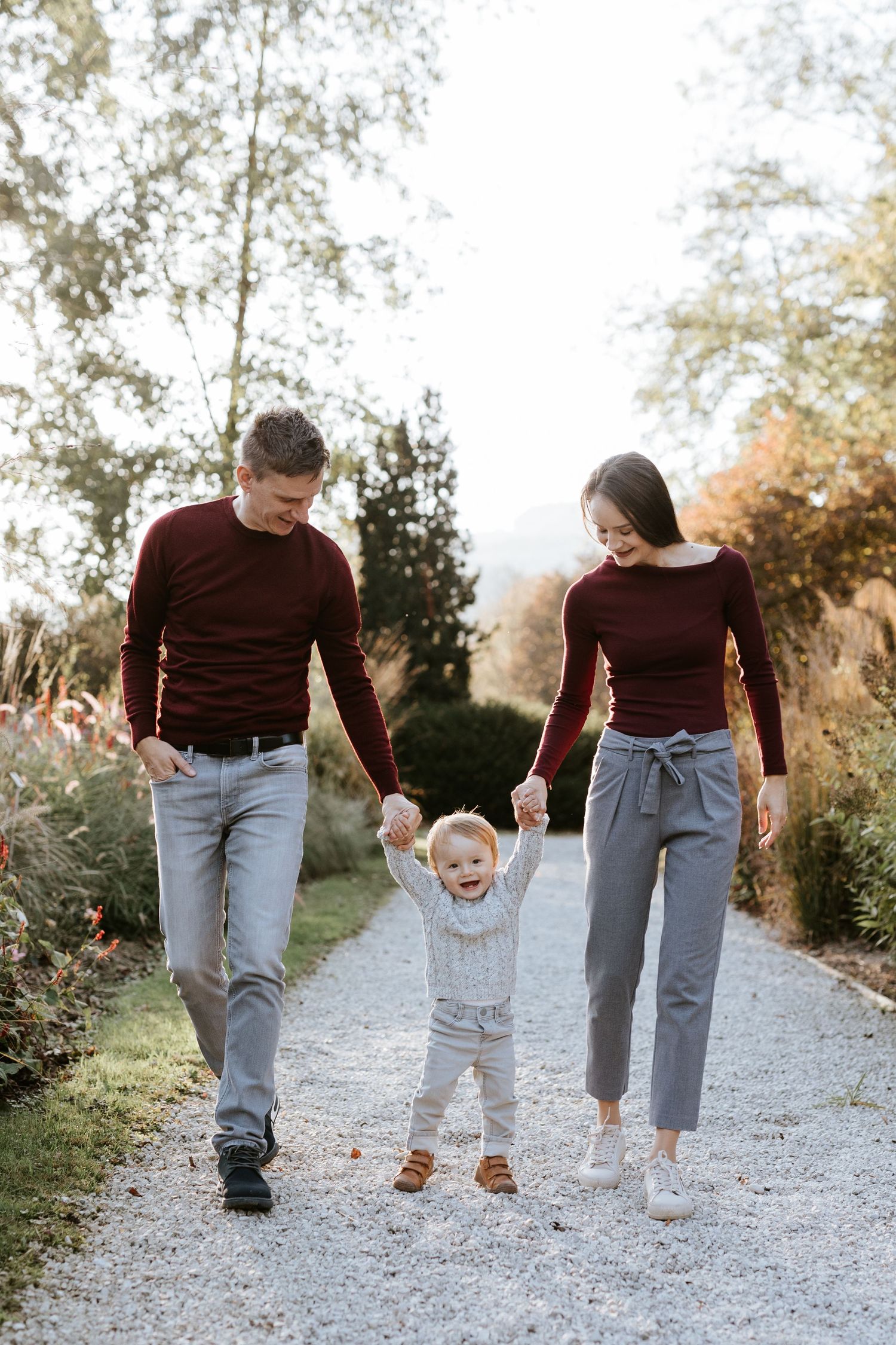 Young family with toddler walking hand-in-hand on garden path in autumn, parents wearing matching burgundy sweaters.