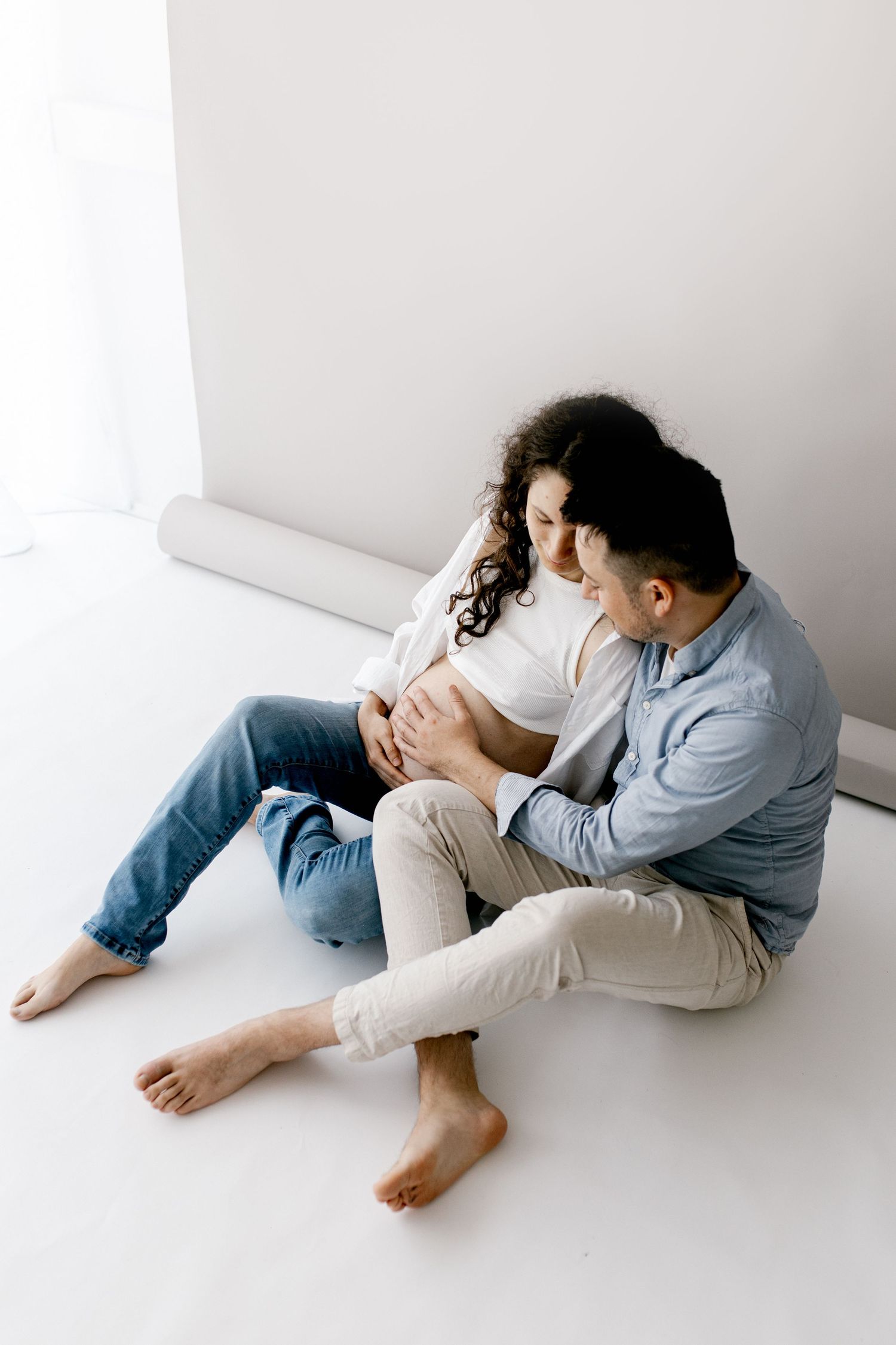 Couple sitting on floor, man tenderly touching pregnant woman's belly in bright white studio.