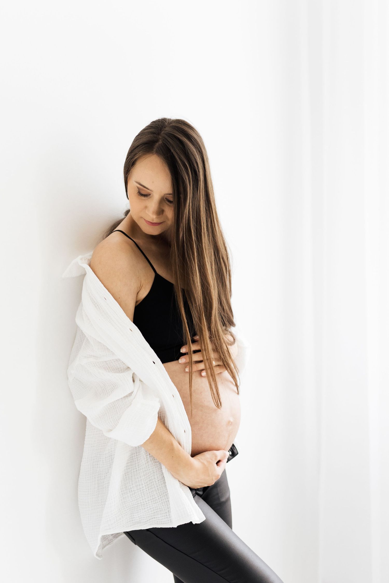 Pregnant woman in white shirt and black top cradling bare baby bump indoors.