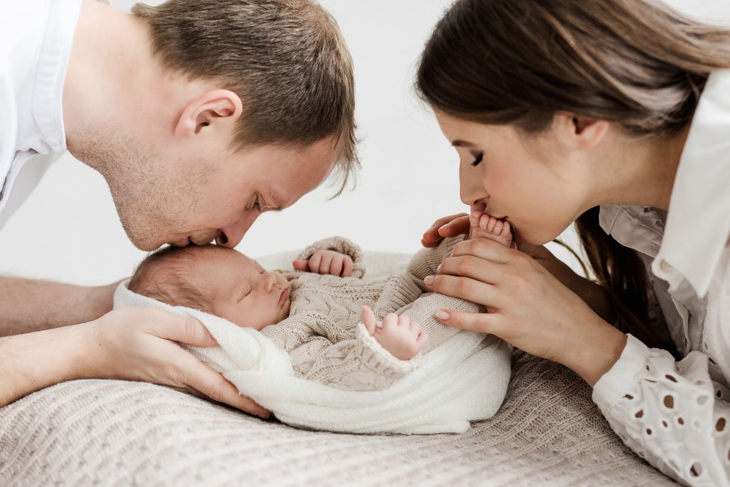 New parents lovingly kissing their newborn baby together in a tender family portrait.
