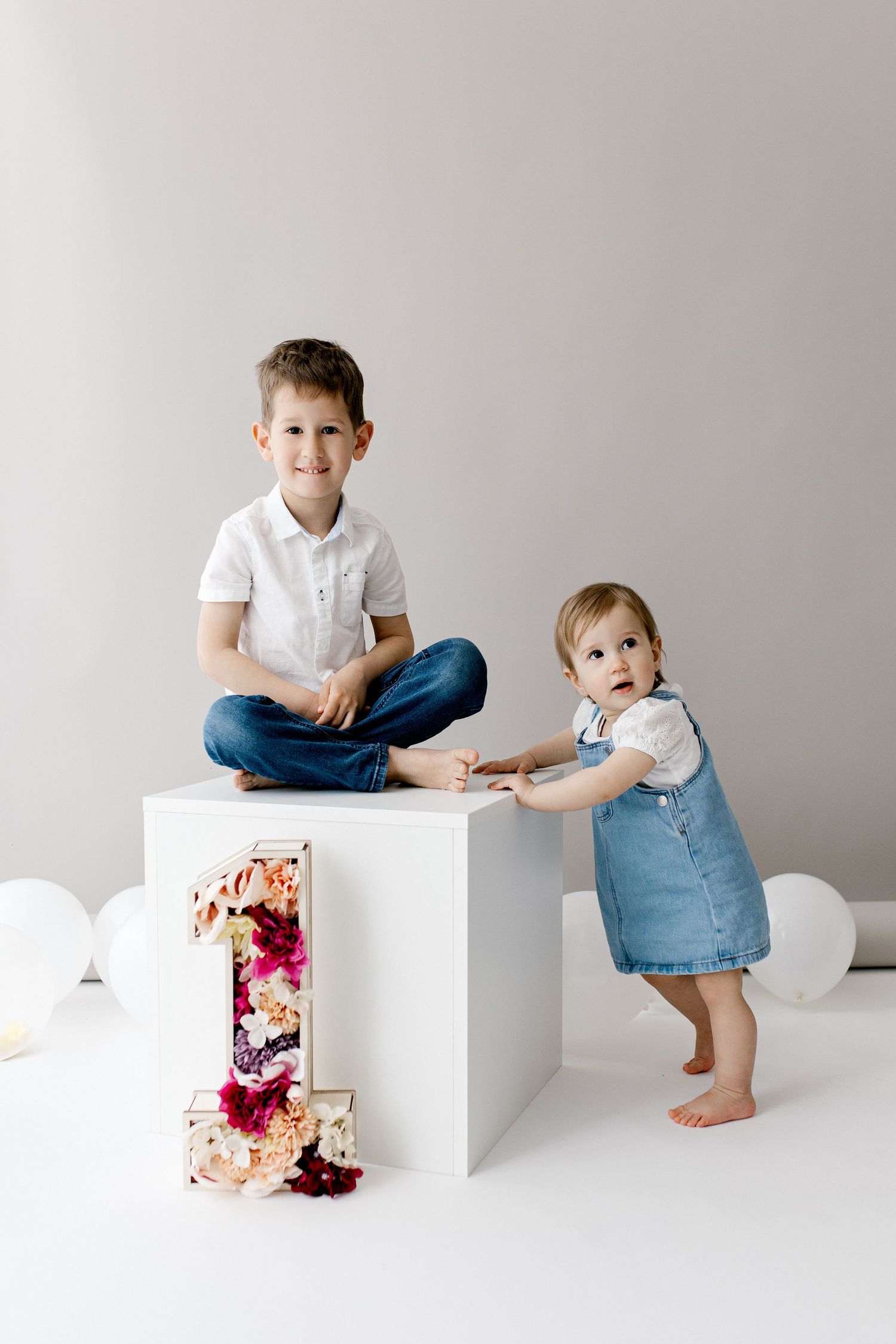 Two young siblings pose with floral number one decoration and white balloons for first birthday celebration.