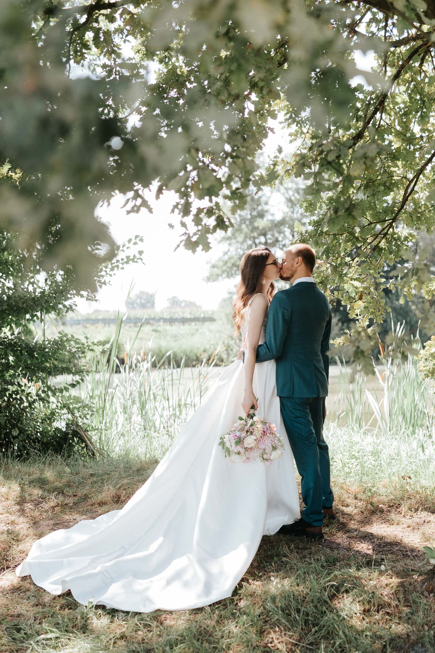 Bride and groom share a kiss under a canopy of green trees beside a serene meadow.
