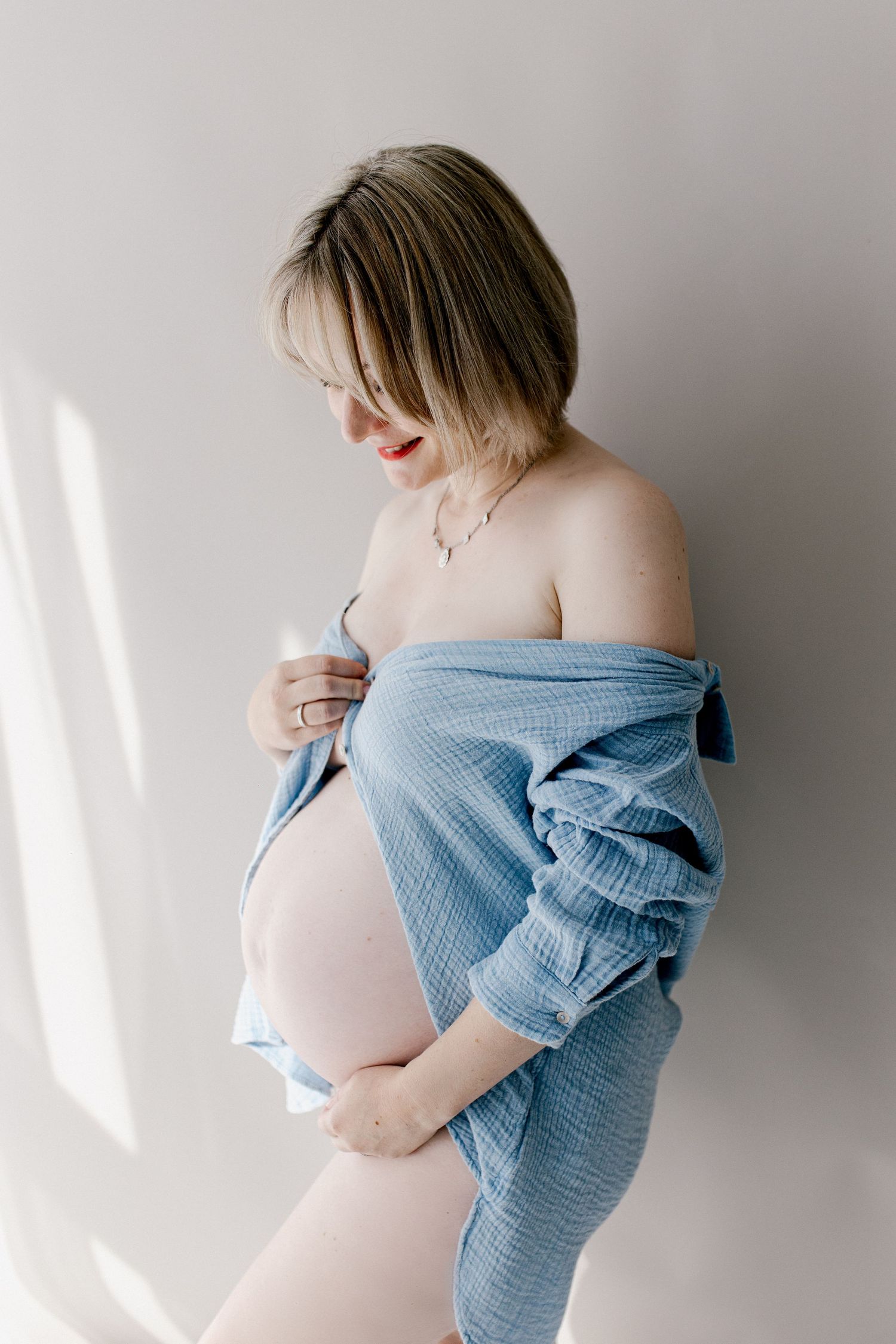 Pregnant woman in blue muslin shirt holding baby bump against white wall.