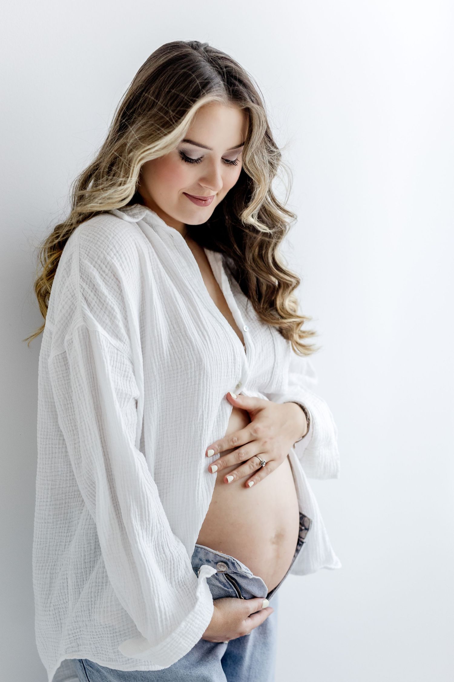 Elegant pregnant woman in white shirt and jeans gently holding her bare baby bump.