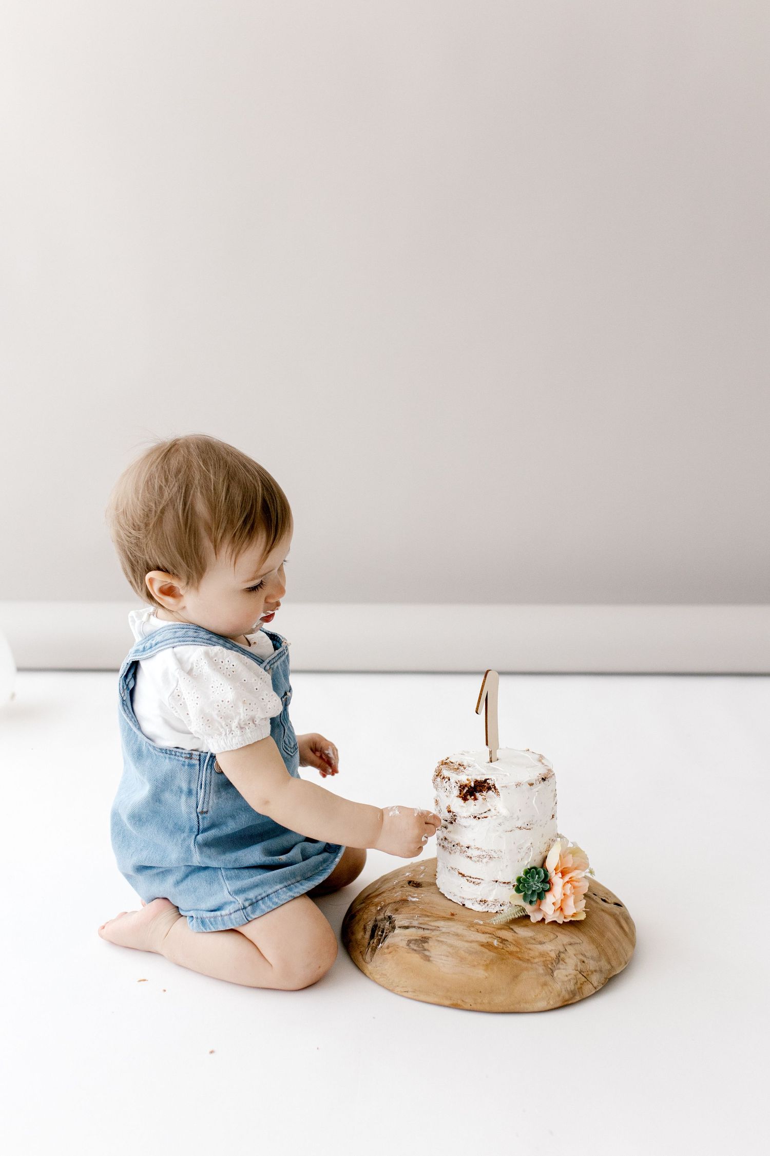 An adorable baby in denim overalls reaches toward a small white first birthday smash cake.