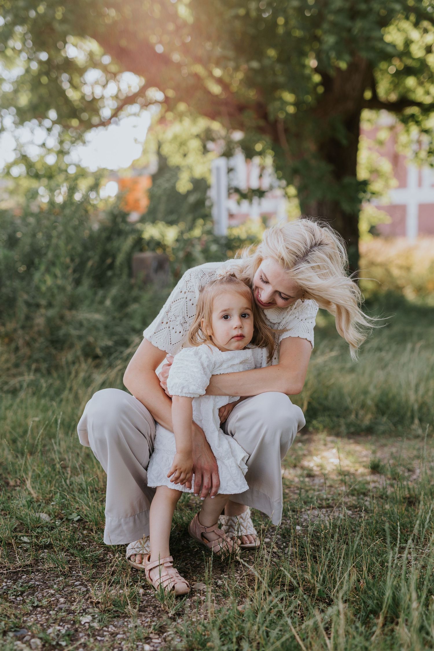 Blonde mother crouching to hug young daughter in white dress in golden sunlit garden near red building.