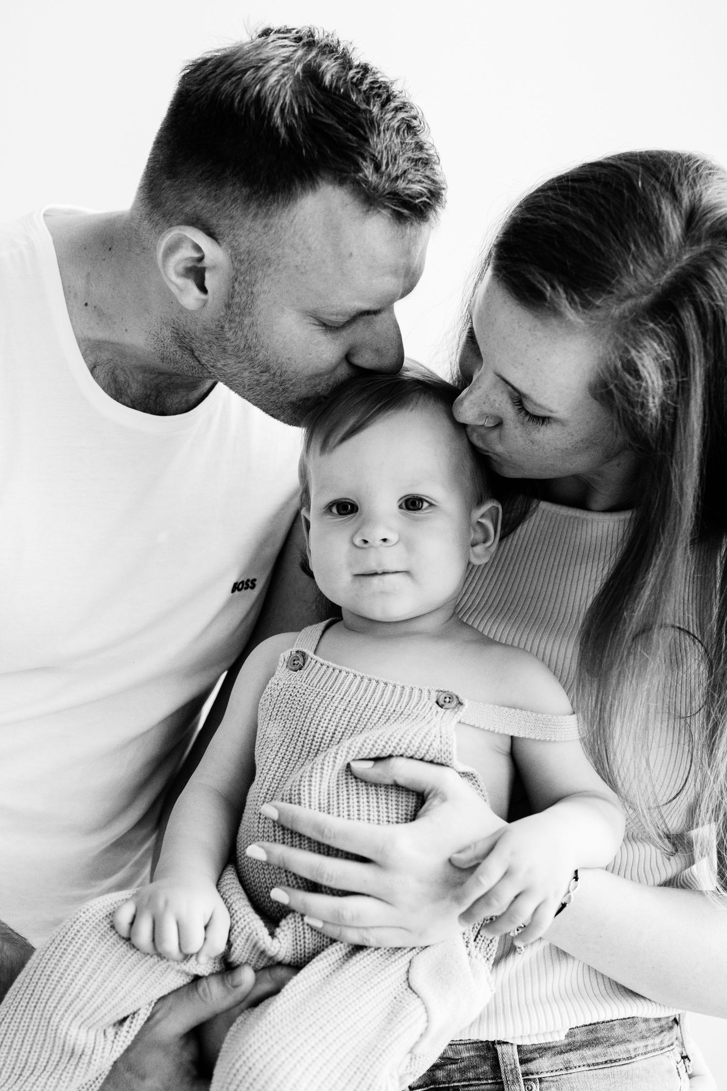 Black and white portrait of two parents lovingly kissing their baby on the cheeks in a studio setting.