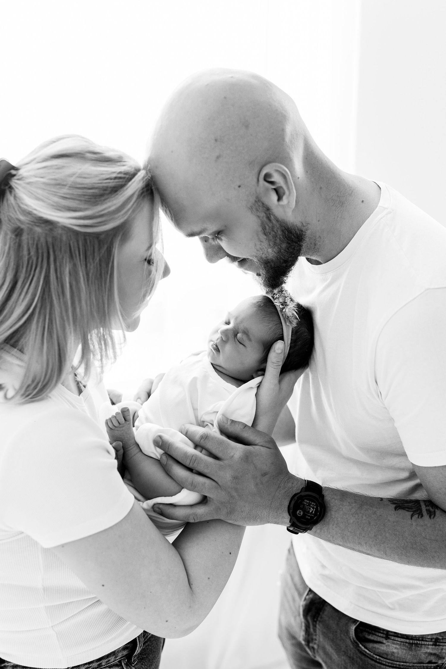 Black and white photo of parents touching foreheads while holding their sleeping newborn baby.
