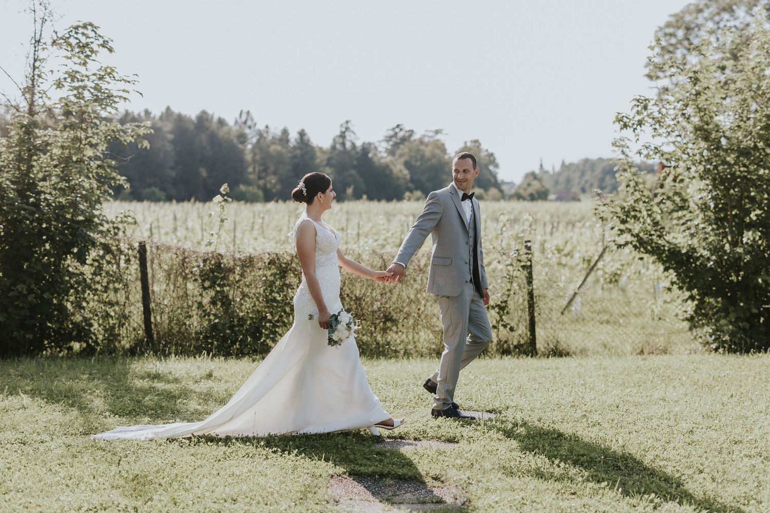 A bride and groom hold hands walking along a garden path with lush green fields and trees in the background.
