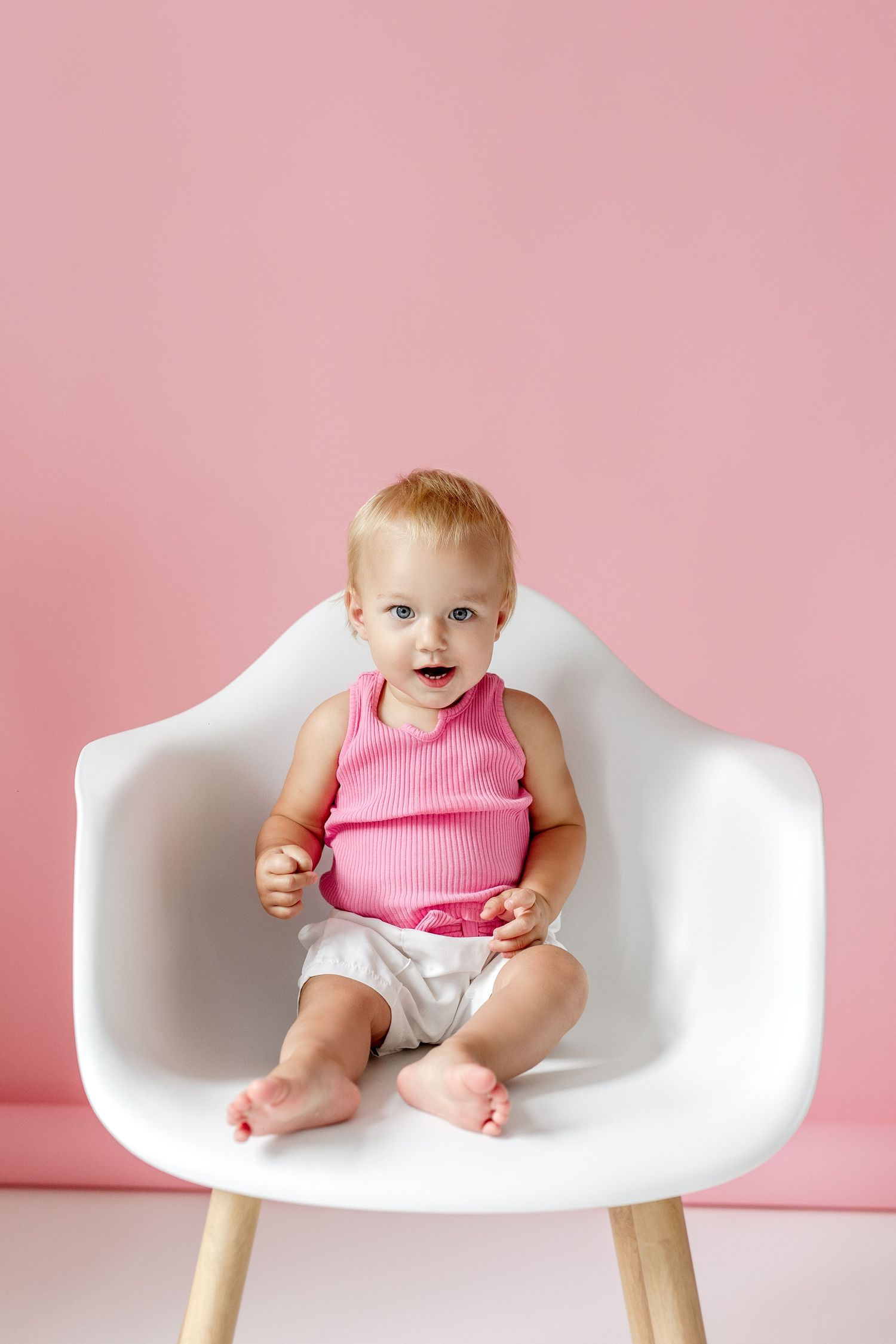 Cute baby in pink top sitting in white modern chair against pink background.