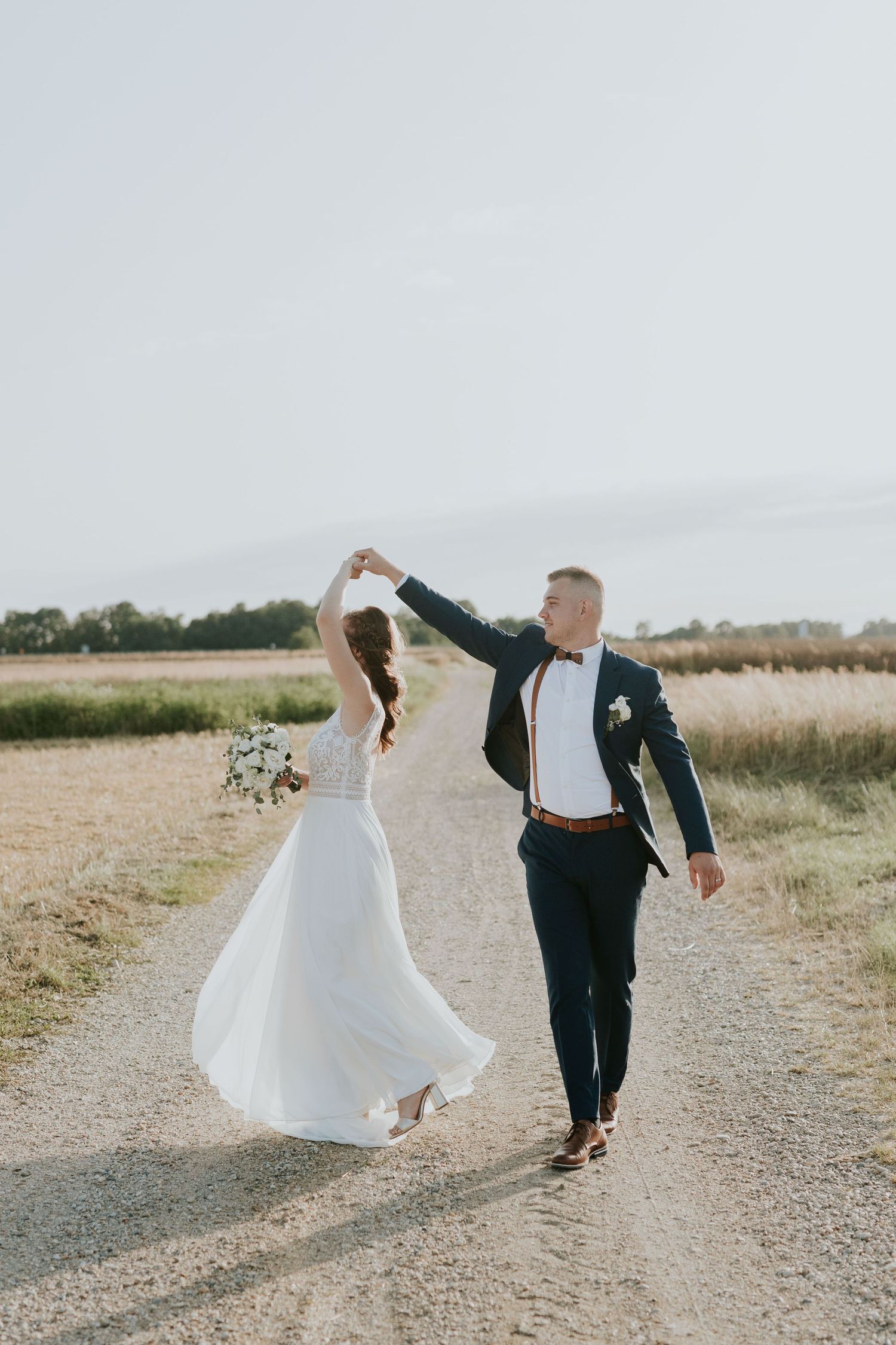 Joyful bride and groom dancing on a rural dirt path surrounded by open fields on a sunny day.
