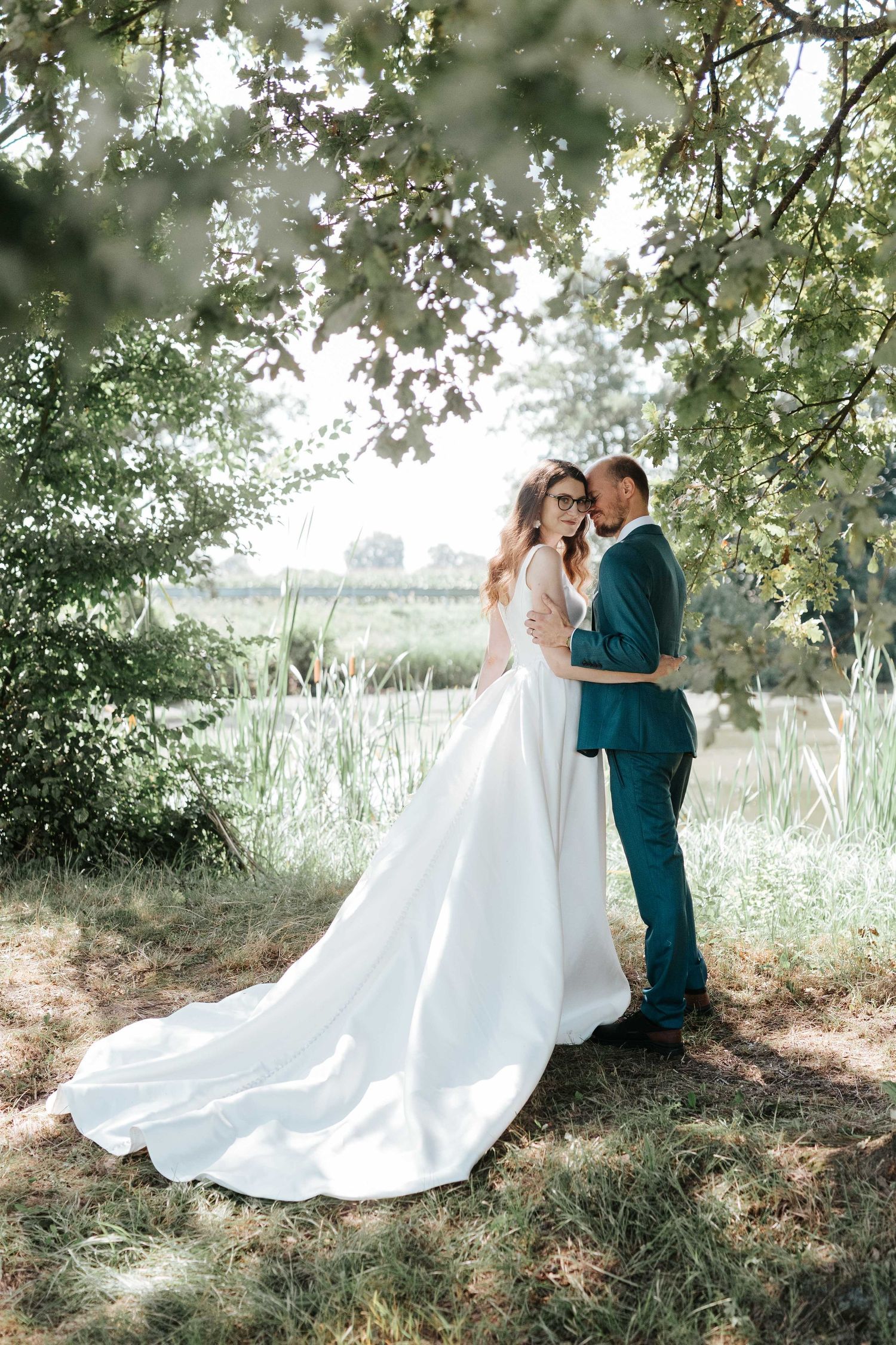 Bride and groom embrace romantically under lush green tree canopy in a sunlit meadow.