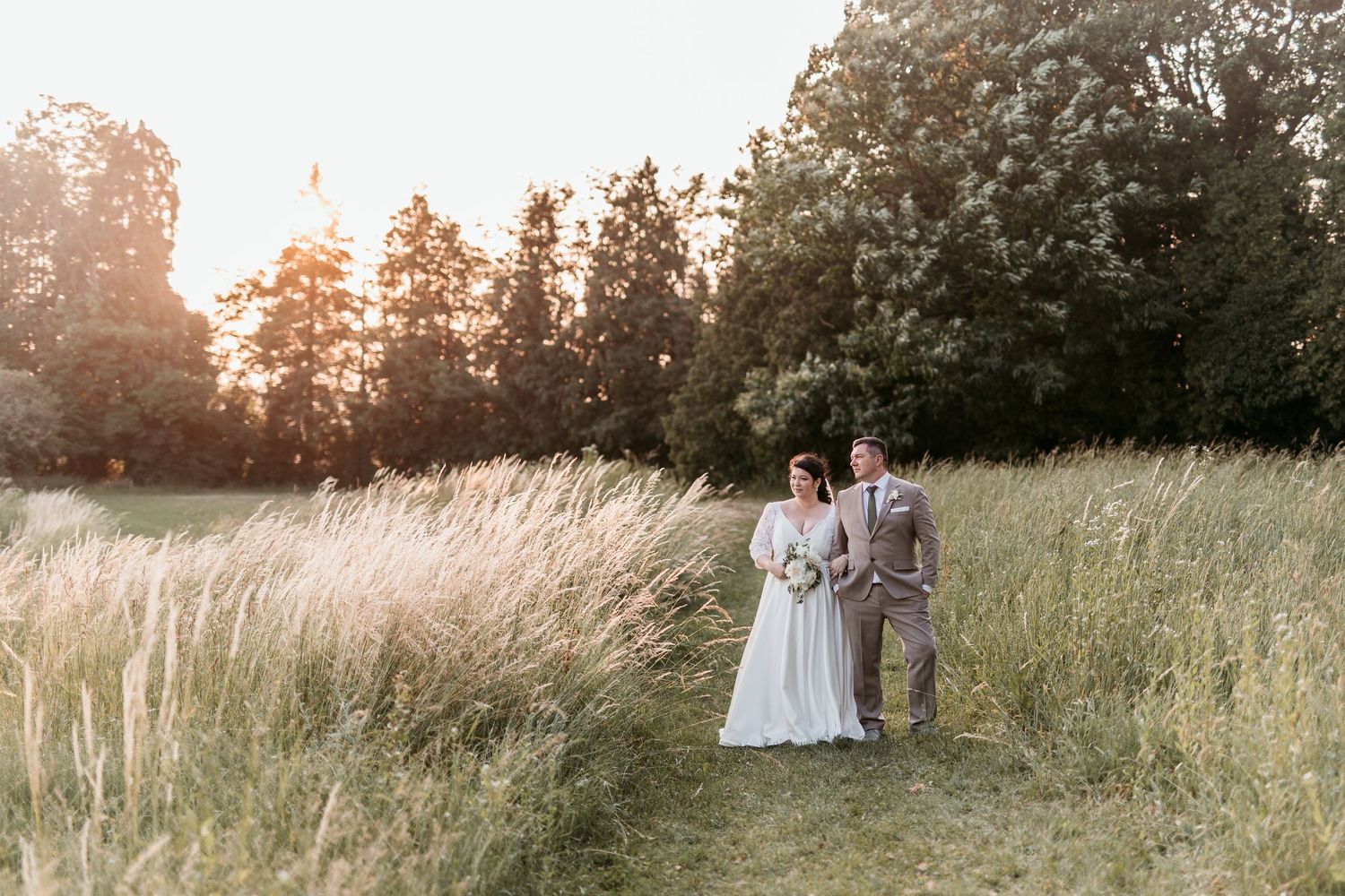 Bride and groom standing in golden sunlit meadow surrounded by tall grass and trees.