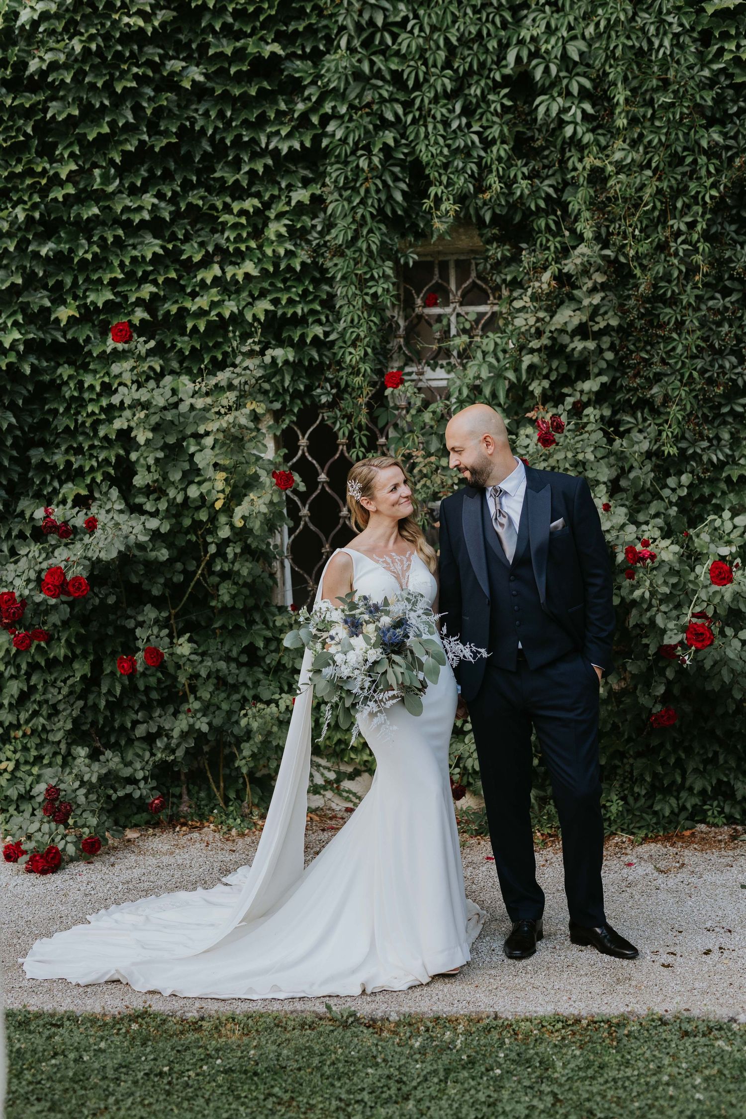 Bride in lace gown and groom in navy suit pose before an ivy-covered wall with red roses.