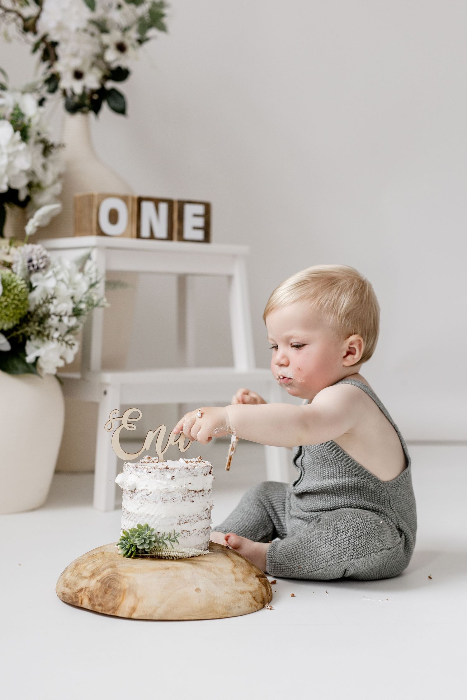 Baby in gray romper poking first birthday cake with name topper and ONE blocks.