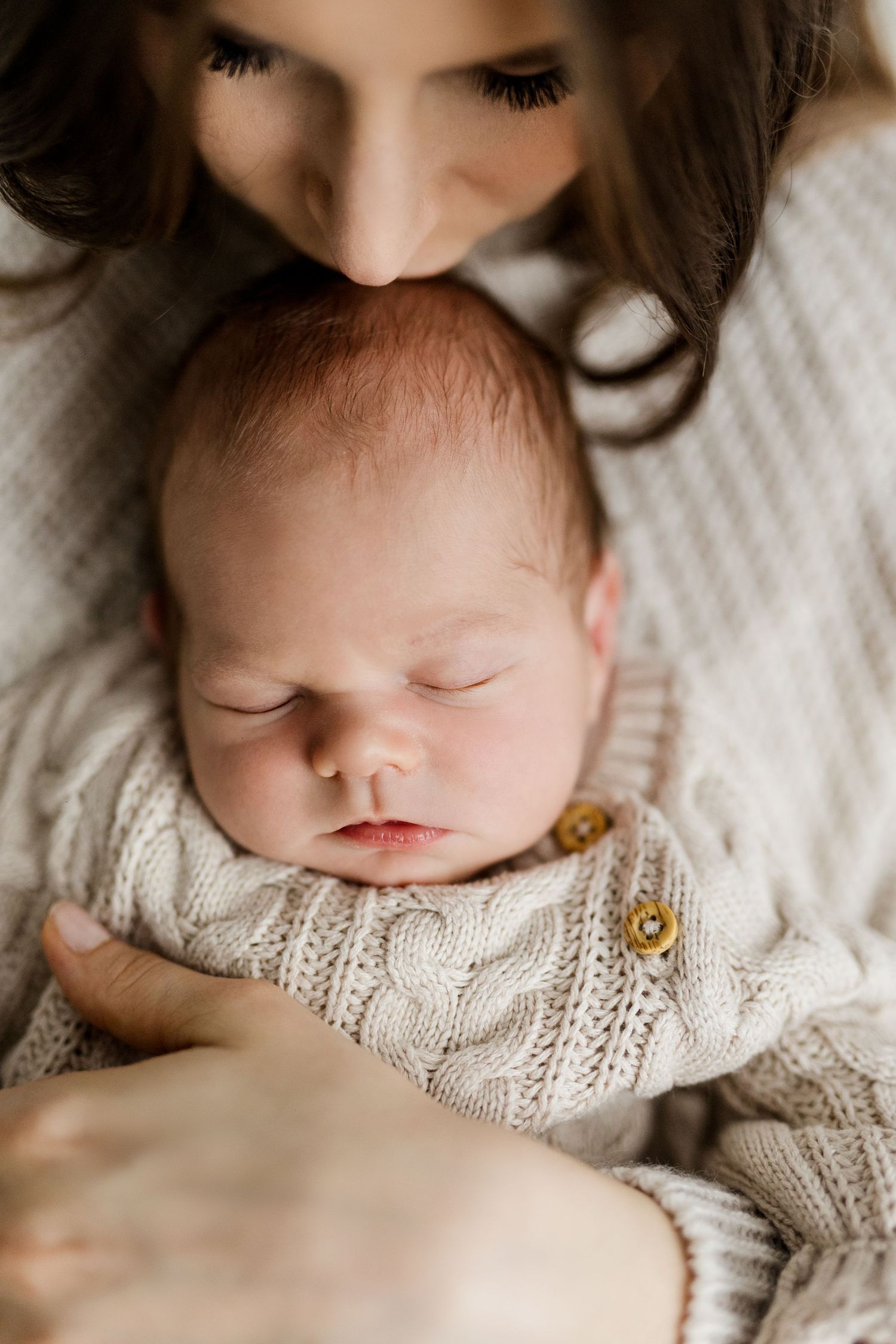 Mother gently kissing her sleeping newborn baby dressed in a cozy cream cable-knit outfit.